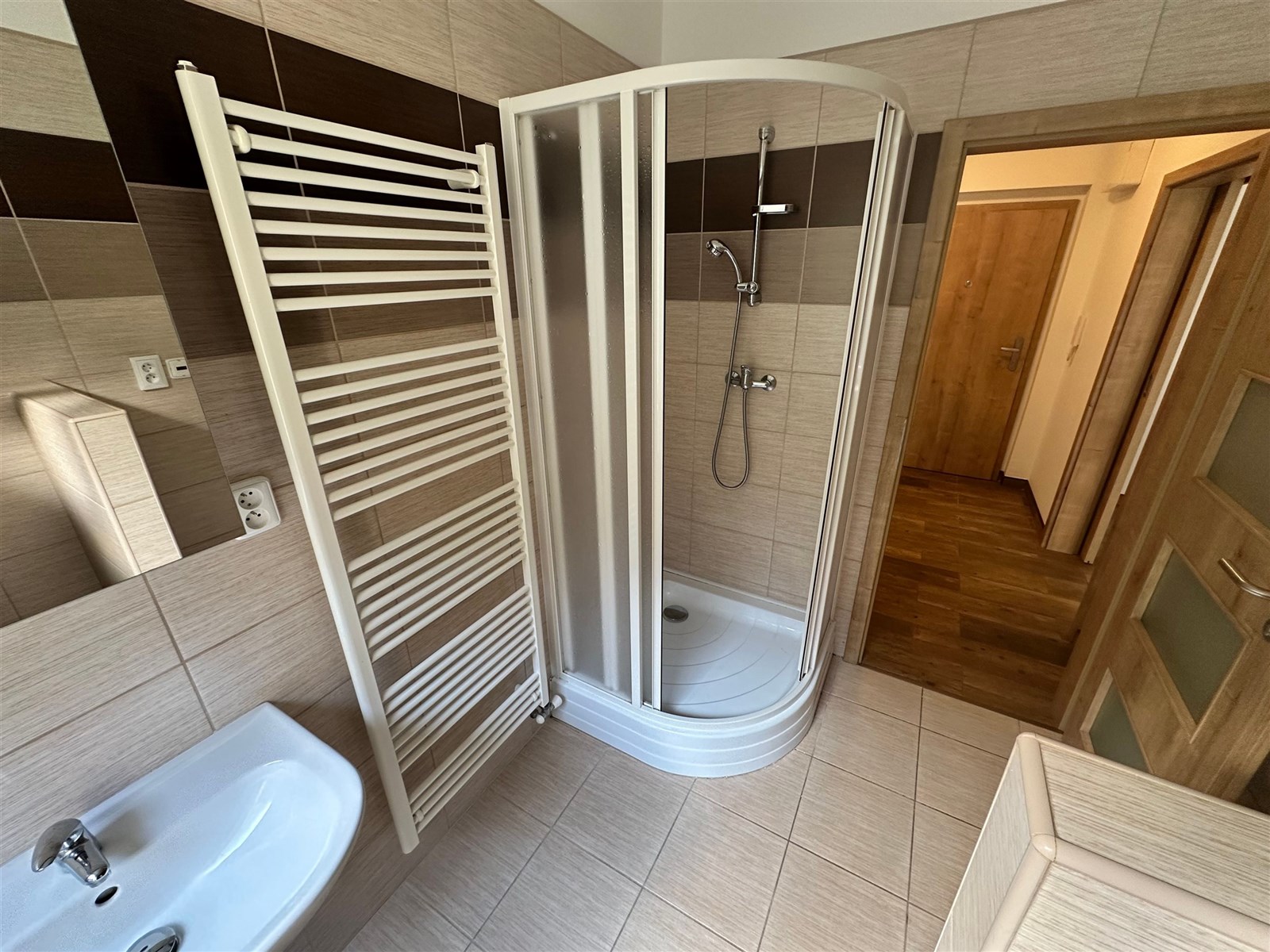 Moderní koupelna s dlažbou, sprchovým koutem a žebřinovým radiátorem, světlé obklady a dřevěné dveře vytváří elegantní vzhled.