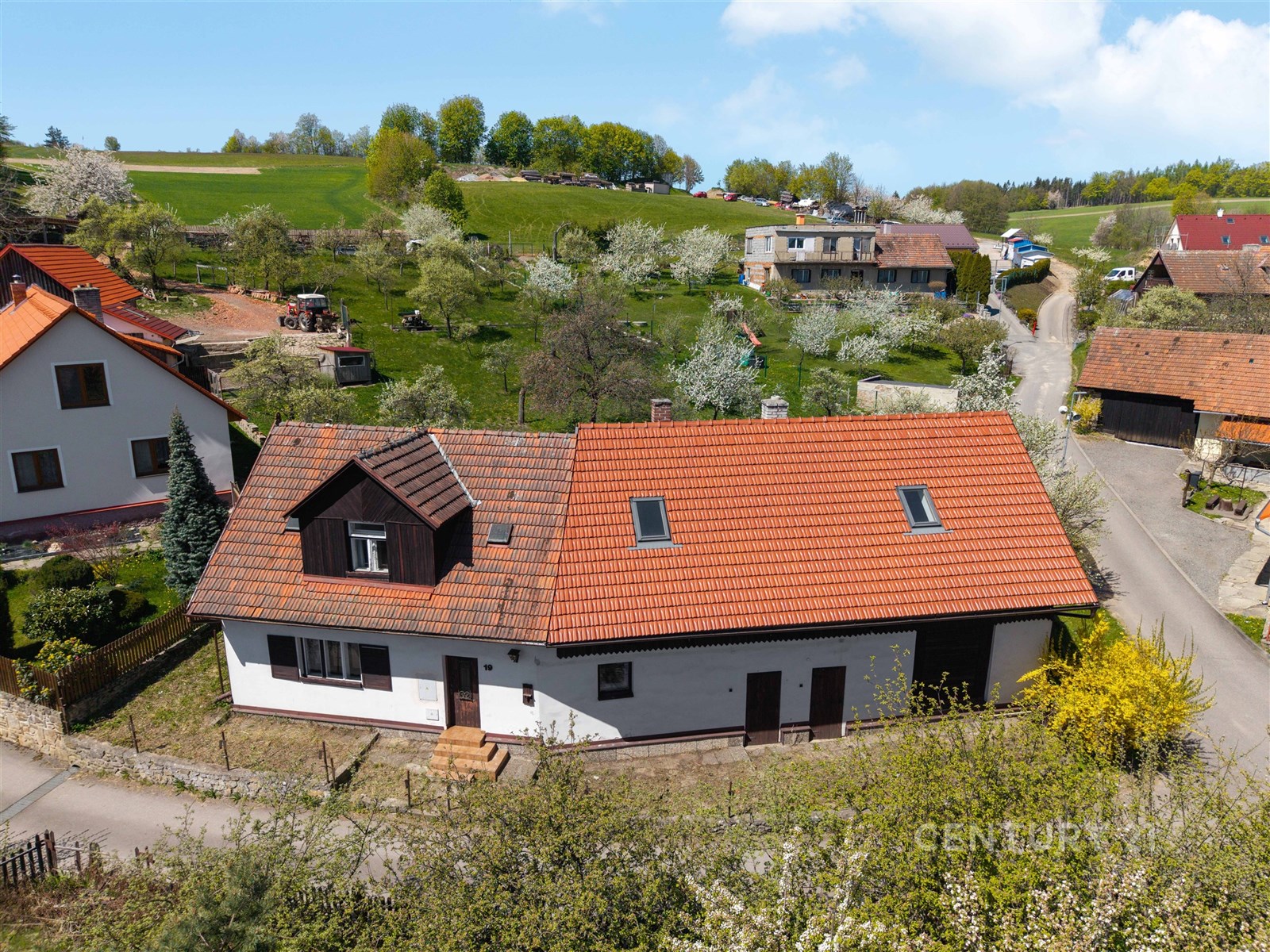 Tradiční venkovský dům s červenou taškovou střechou a světlou fasádou, obklopený zelení a udržovanými zahradami, v klidné ulici.