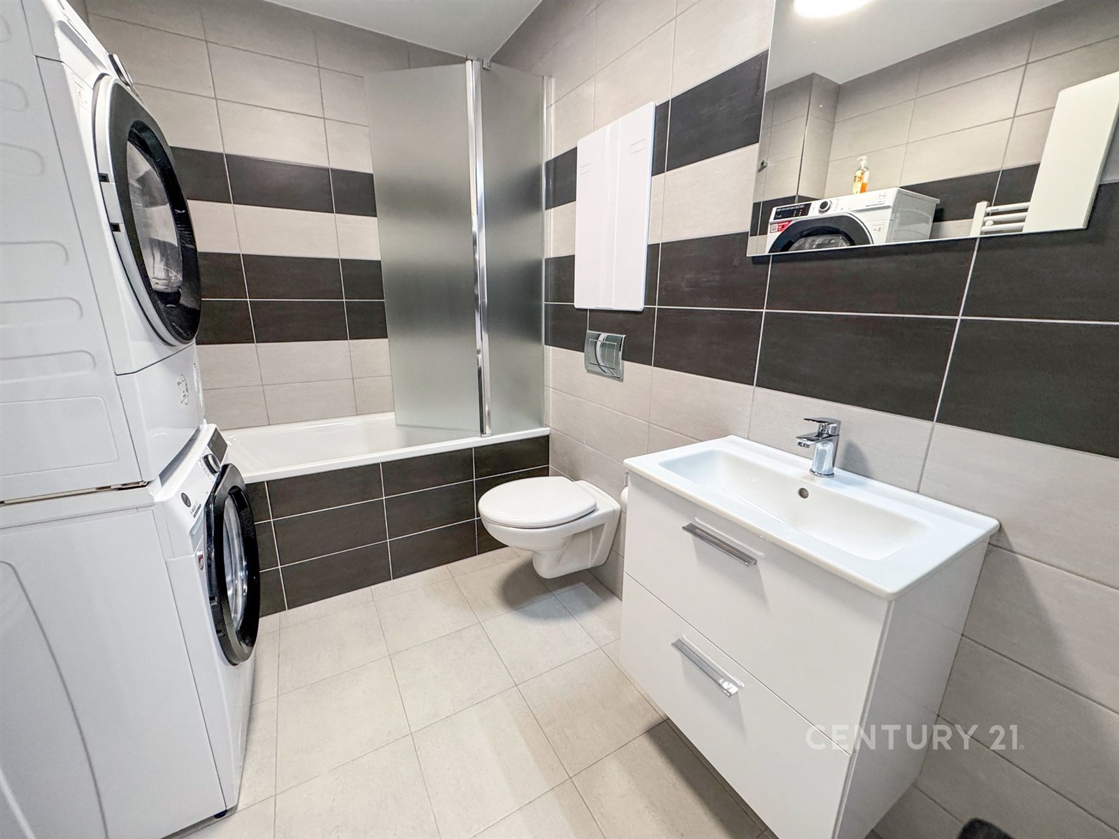 Moderní koupelna s dlažbou, vestavěným umyvadlem a úložnou skříňkou. Kombinace světlých a tmavých obkladů, pračka se sušičkou.