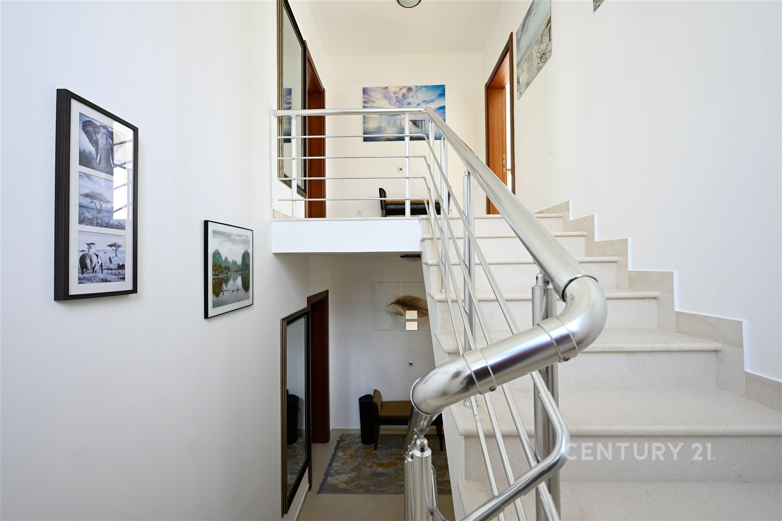 Schodiště s nerezovým zábradlím, moderní design, světlé stěny, dekorativní obrazy, prostorný interiér, kvalitní povrchová úprava, dřevěné prvky.