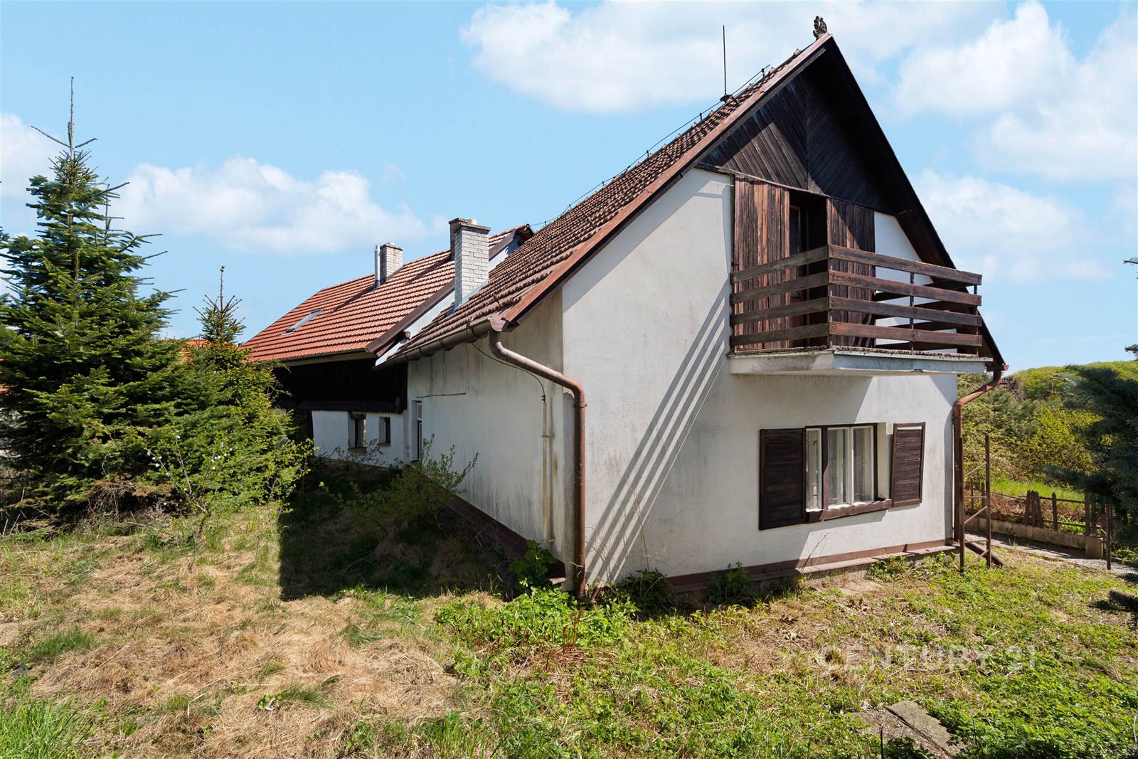 Exteriér rodinného domu se šikmou střechou, dřevěným balkonem a okny s okenicemi, nacházející se v klidné oblasti obklopené zelení.