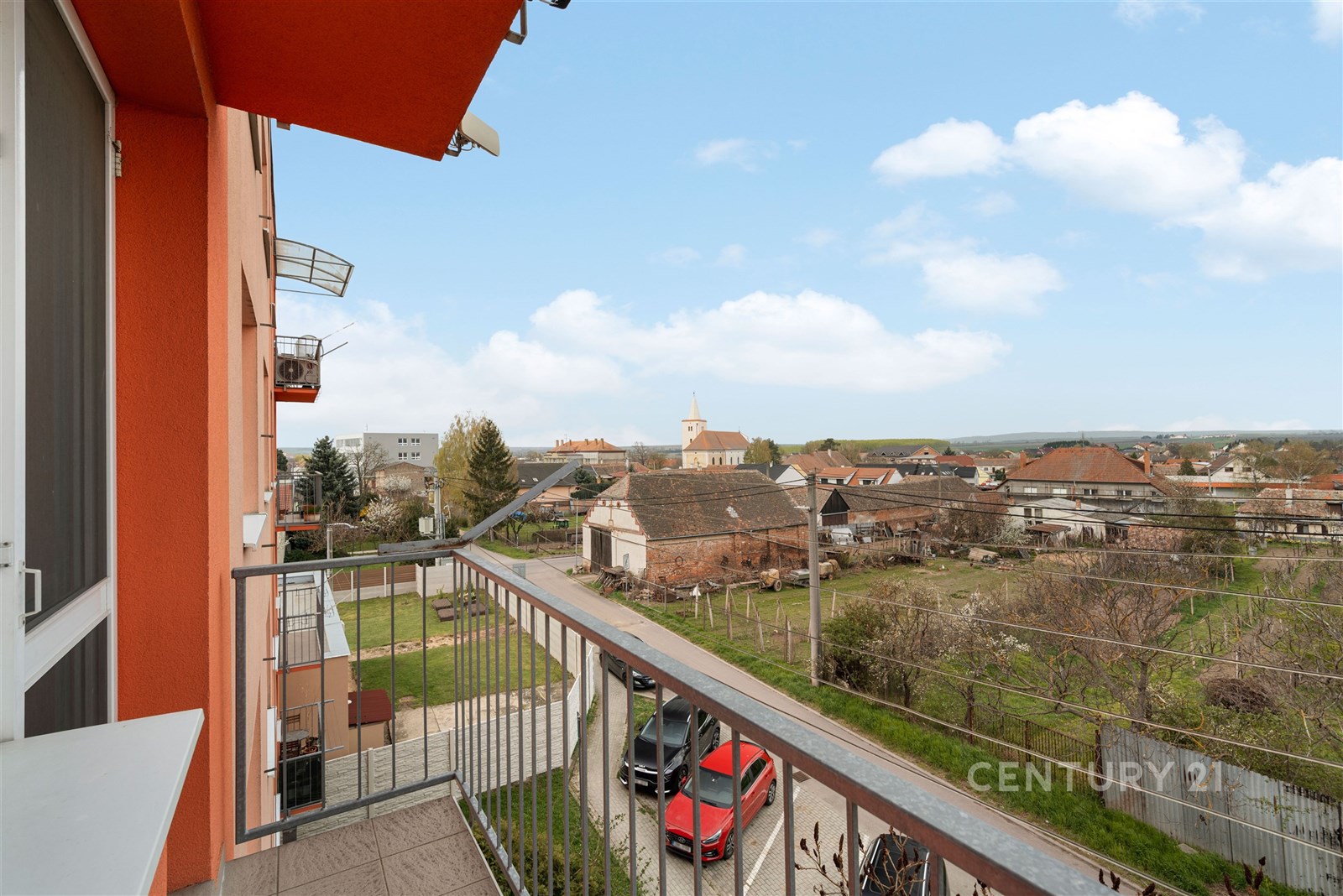 Balkon s výhledem na okolní zástavbu, kovové zábradlí a dlažba, světlé venkovní prostředí, vhodný pro relaxaci na čerstvém vzduchu.