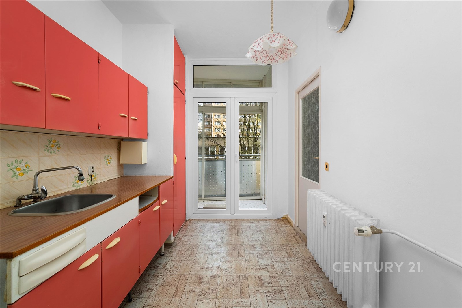 Kuchyň s parketovou podlahou a červenými skříňkami, světlý prostor s velkými okny a radiátorem, rekonstrukce vhodná.