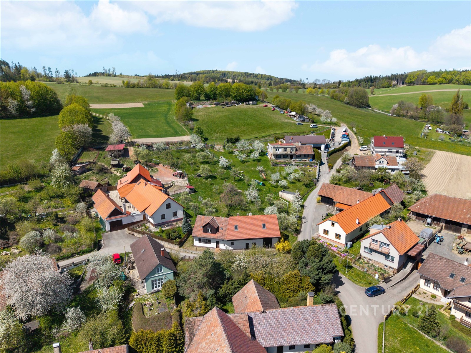 Letecký pohled na malebnou vesnici s rodinnými domy, zahradami a okolní krajinou, nabízící klidné prostředí a přírodní scenérie pro bydlení.