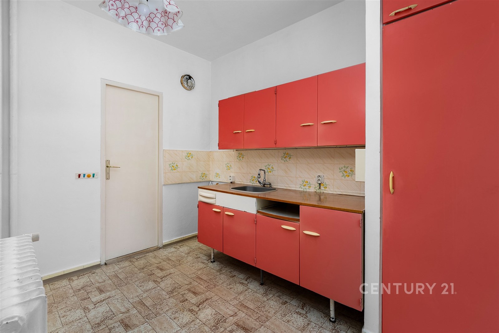 Kuchyň s retro skříňkami, dlažbou a bílými stěnami. Vhodná k rekonstrukci, nabízí prostory pro kreativní úpravy a osobní styl.