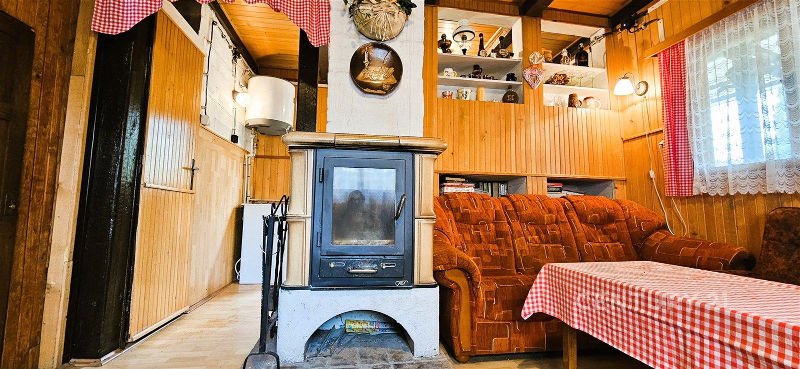 Útulný obývací pokoj s krbovými kamny, dřevěnými obklady a rustikálním nábytkem, textilními závěsy a vestavěnými policemi pro dekorace.