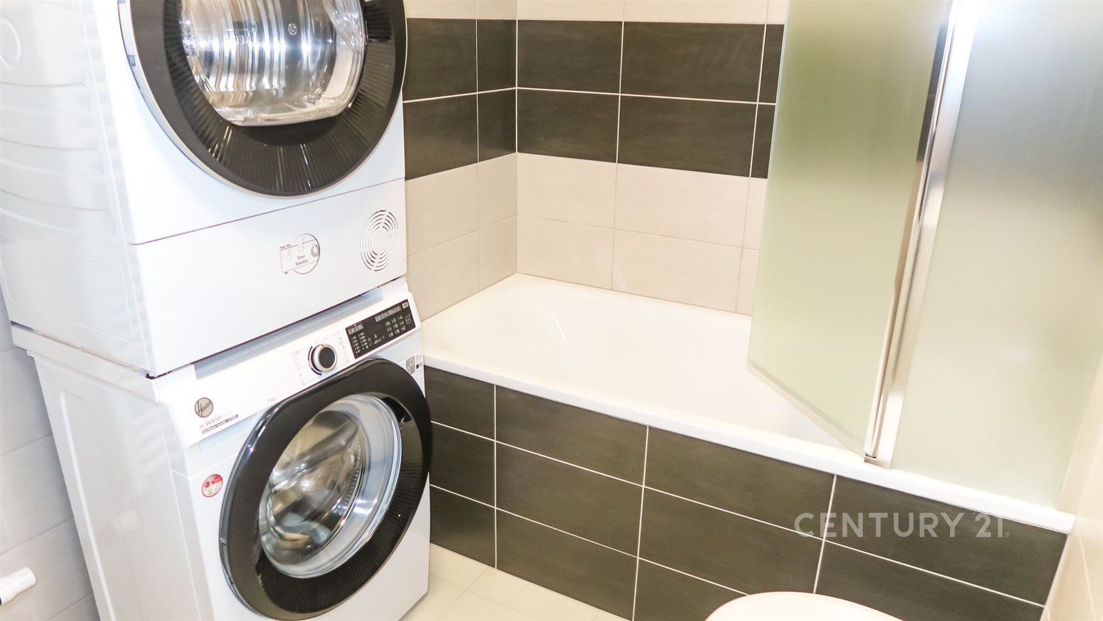 Koupelna s moderními spotřebiči, kombinace pračky a sušičky, keramická dlažba, světlé a tmavé dlaždice, sprchový kout s matným sklem.