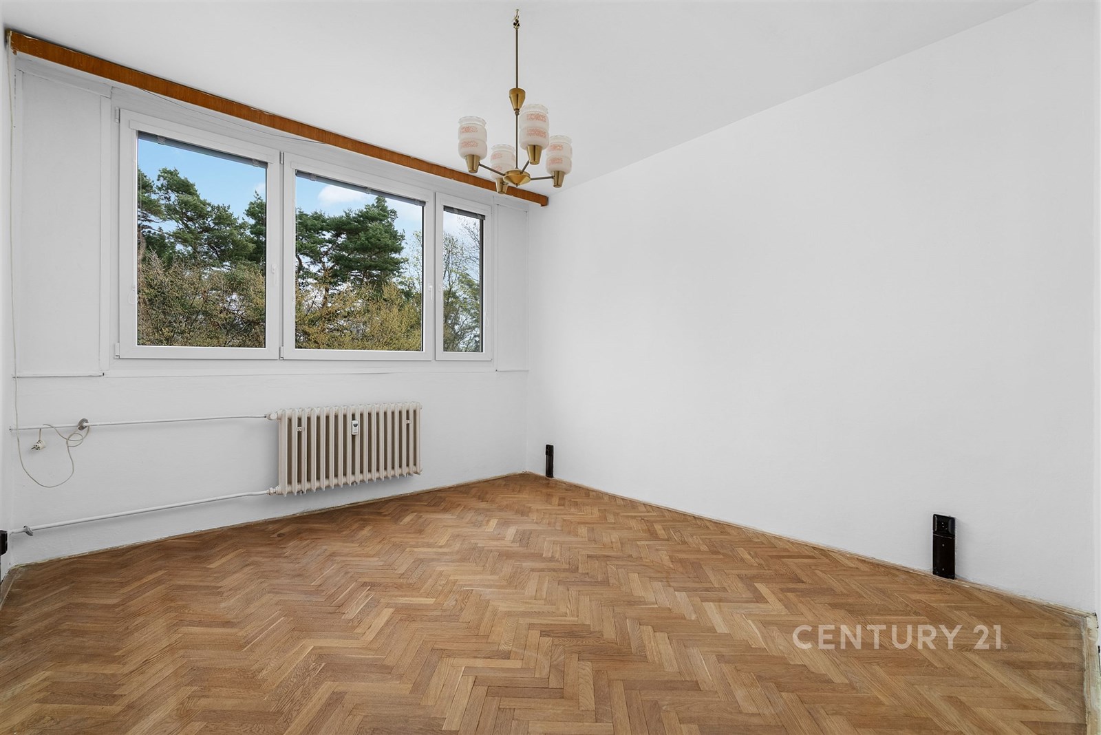 Světlý obývací pokoj s parketovou podlahou a velkými okny, které nabízejí výhled na zeleň, ideální pro moderní úpravu dle vlastního stylu.