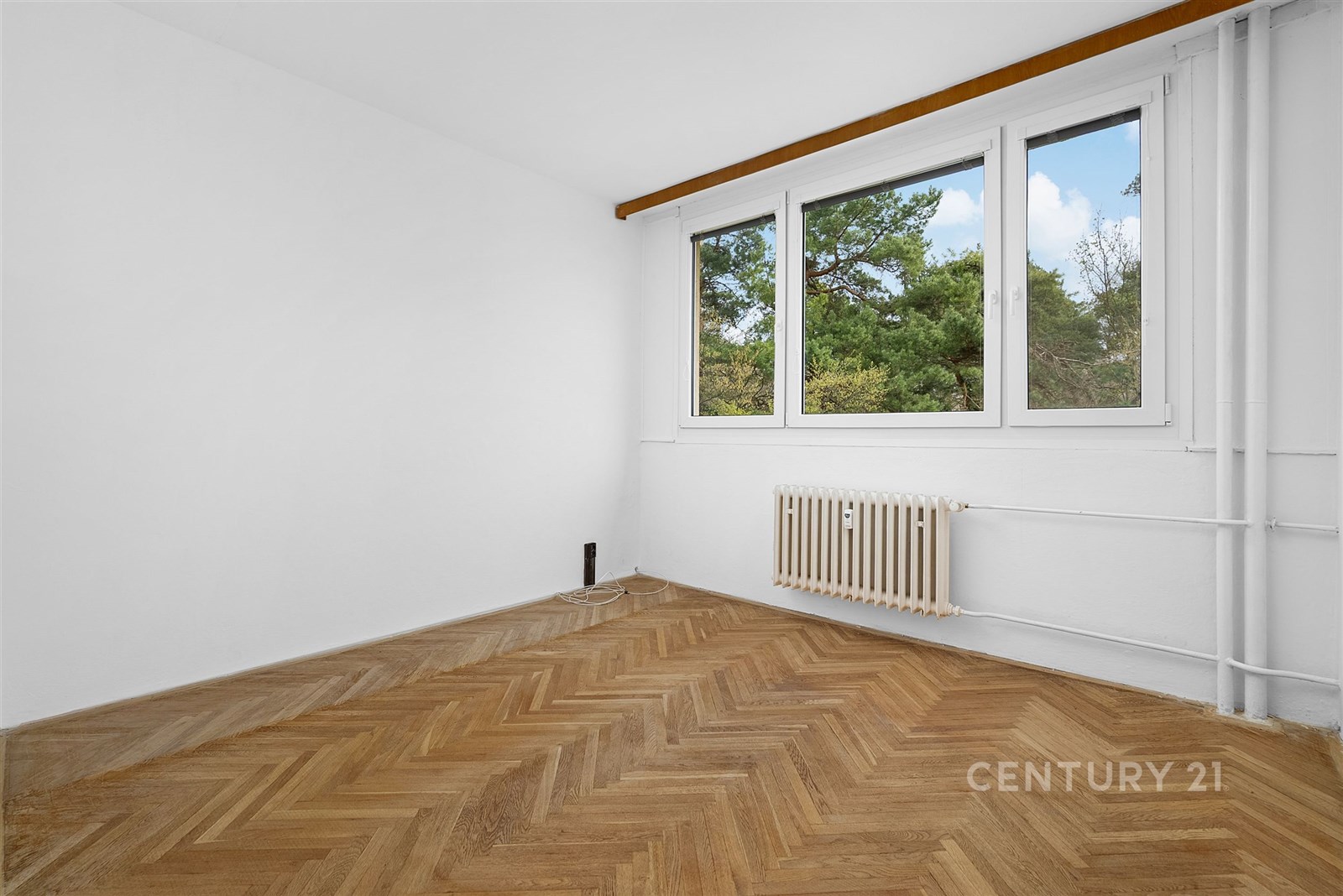 Prázdná místnost s parketovou podlahou, velká okna s výhledem na zeleň, bílé stěny, vytápění radiátorem, světlá a prostorná.