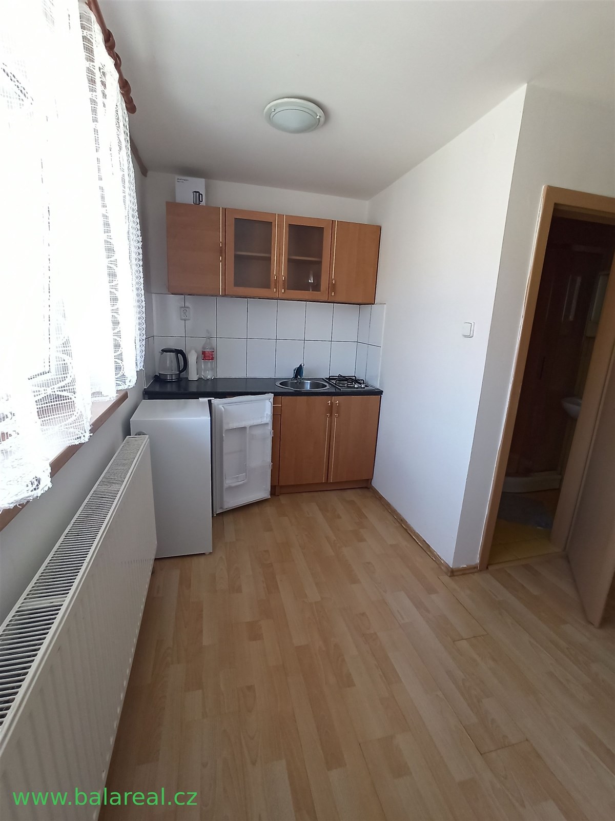 Kompaktní kuchyň s parketovou podlahou, světlé stěny, praktická skříňka a velké okno zajišťující dostatek přirozeného světla.