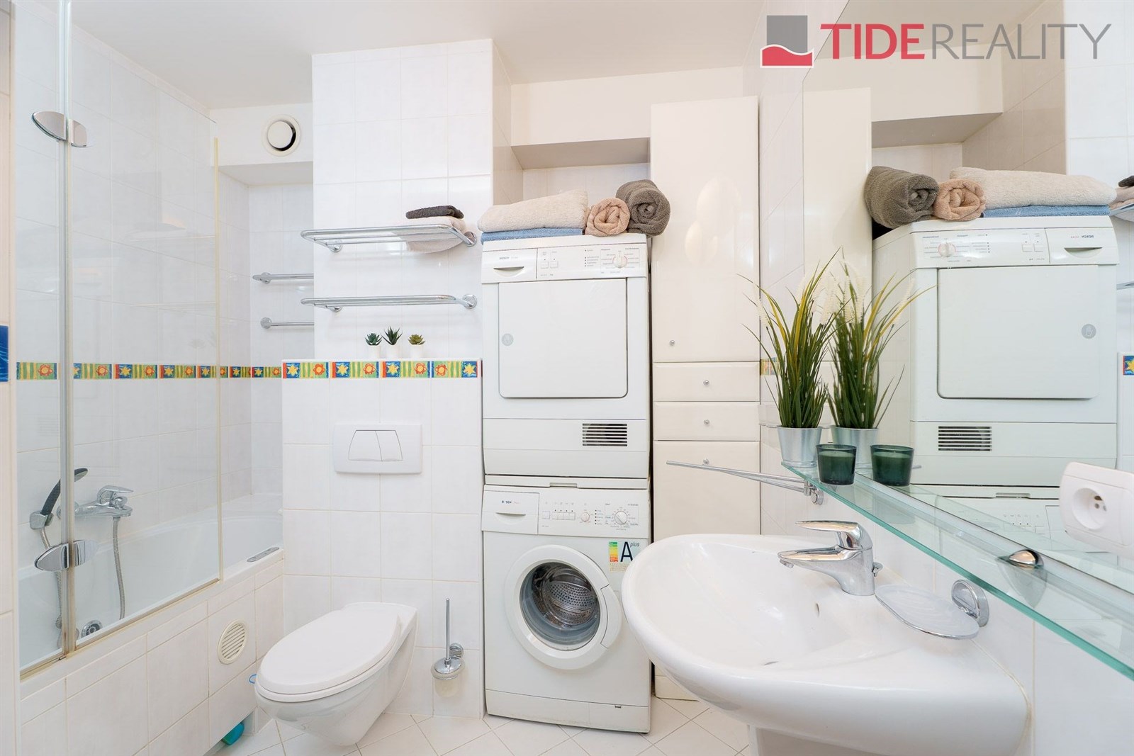 Koupelna s bílými dlaždicemi, vestavěnou skříní a moderní sanitou, obsahující pračku a sušičku pro praktické využití prostoru.