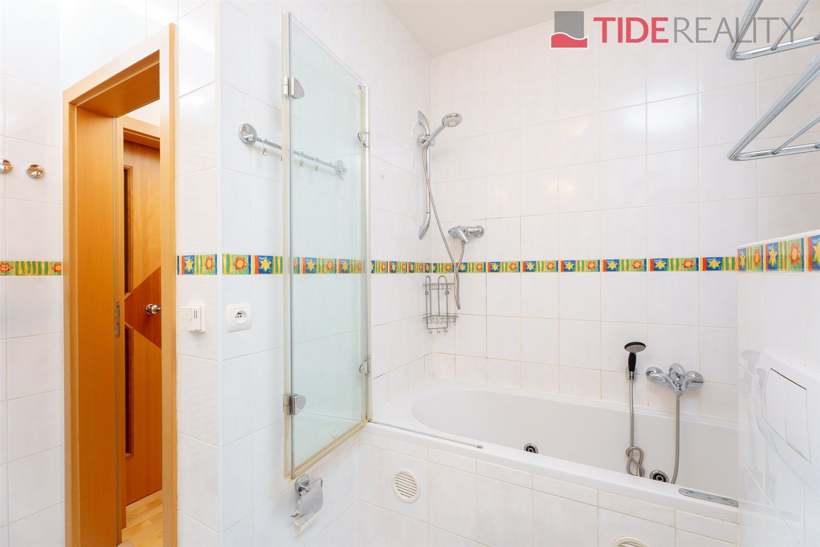 Koupelna s bílými dlaždicemi, moderní vanou a sprchou. Světlý interiér doplňují barevné obklady a dřevěné detaily dveří.