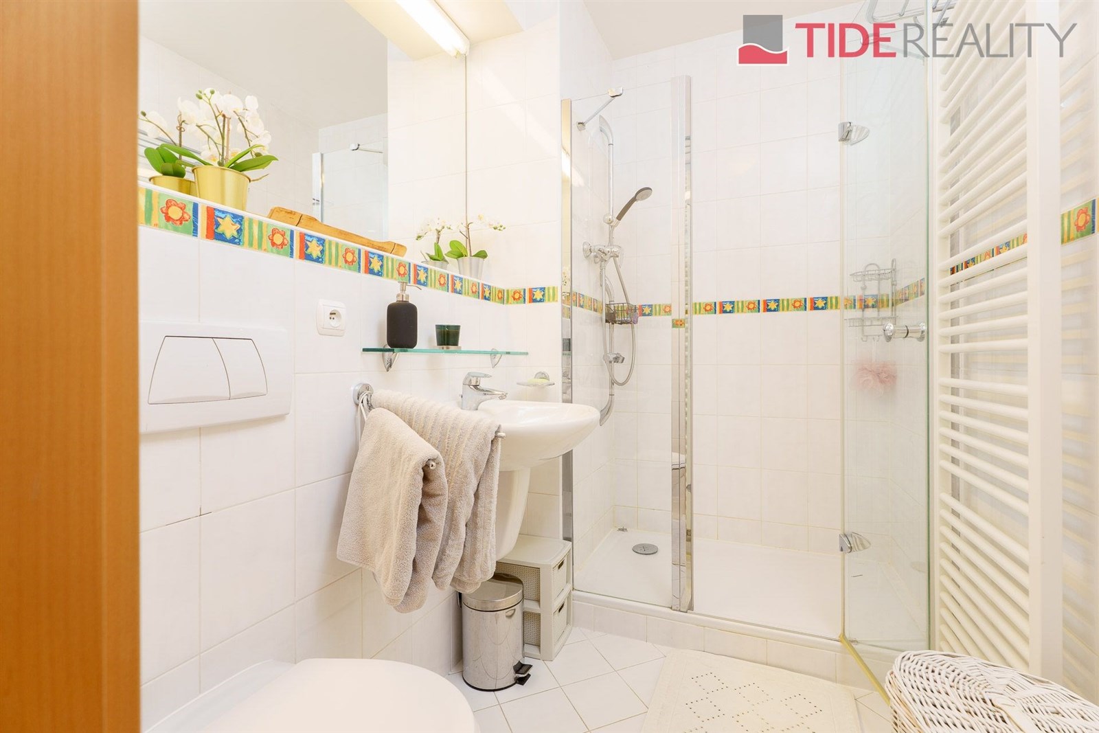 Světlá koupelna s moderními dlaždicemi, sprchovým koutem a nástěnným topením, nabízí praktické uspořádání a čistý styl.