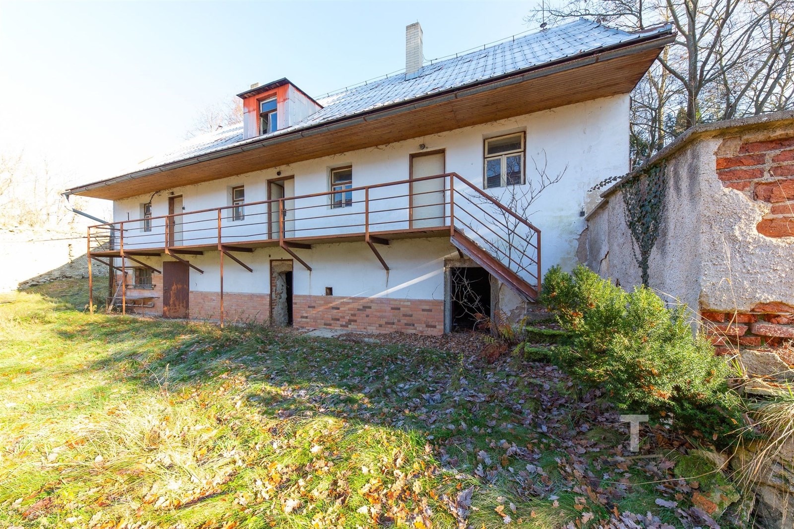 Částečně rekonstruovaný venkovský dům s cihelnou fasádou a dřevěnou terasou, umístěný na klidném pozemku s přírodním okolím.