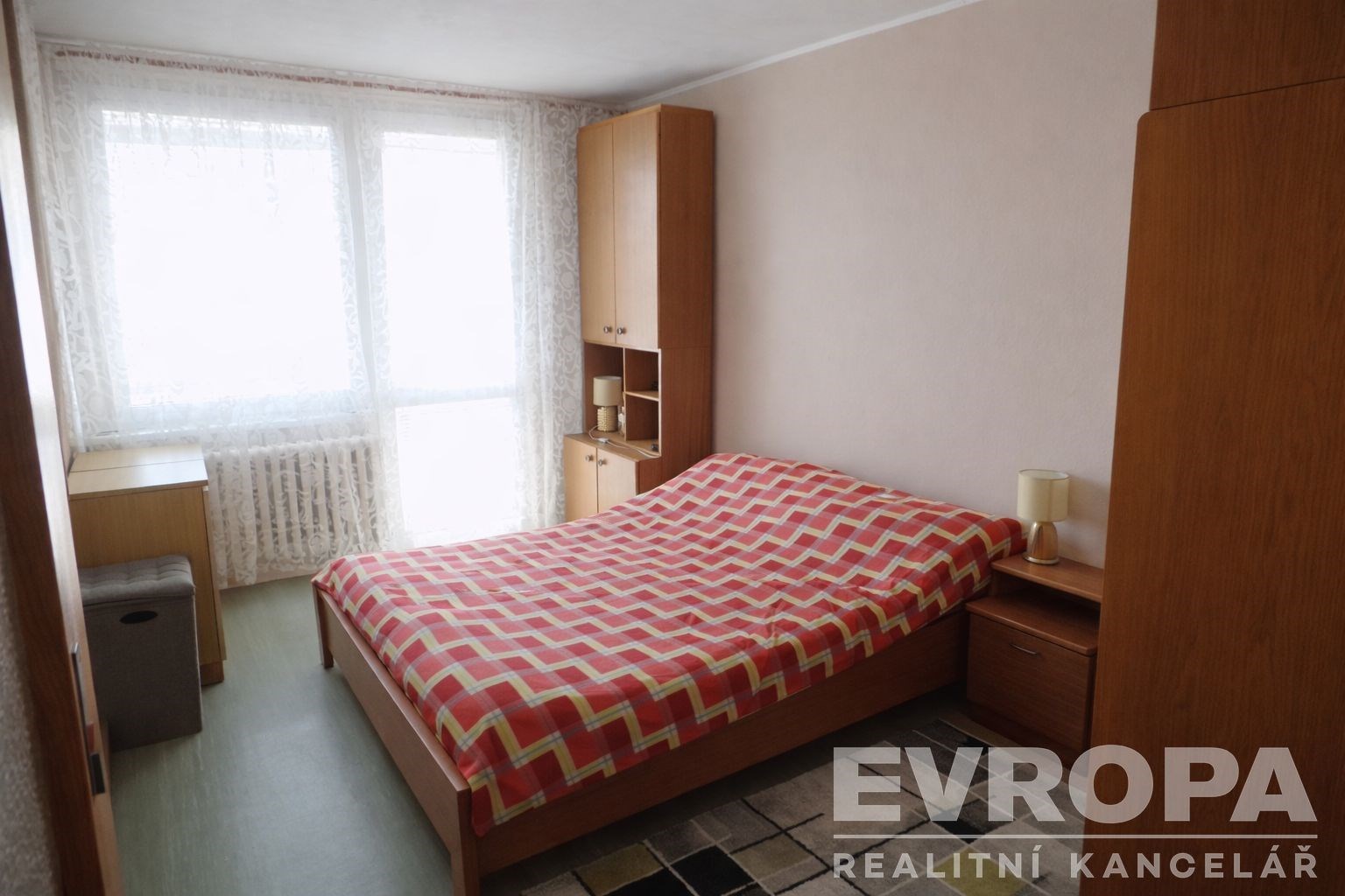 Prostorná ložnice s dřevěným nábytkem, velká okna s průhlednými záclonami, světlá stěna, moderní design, ideální pro komfortní bydlení.