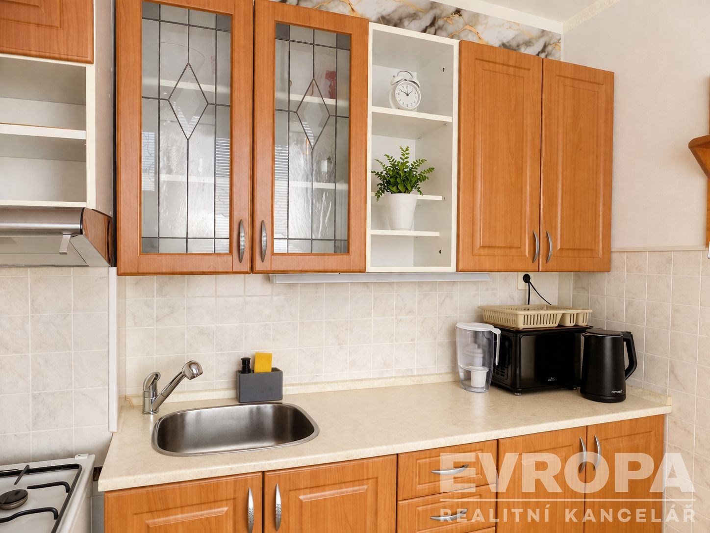 Moderní kuchyň s dřevěnými skříňkami, bílou dlažbou a vestavěnými spotřebiči, poskytuje světlý a praktický prostor pro vaření.