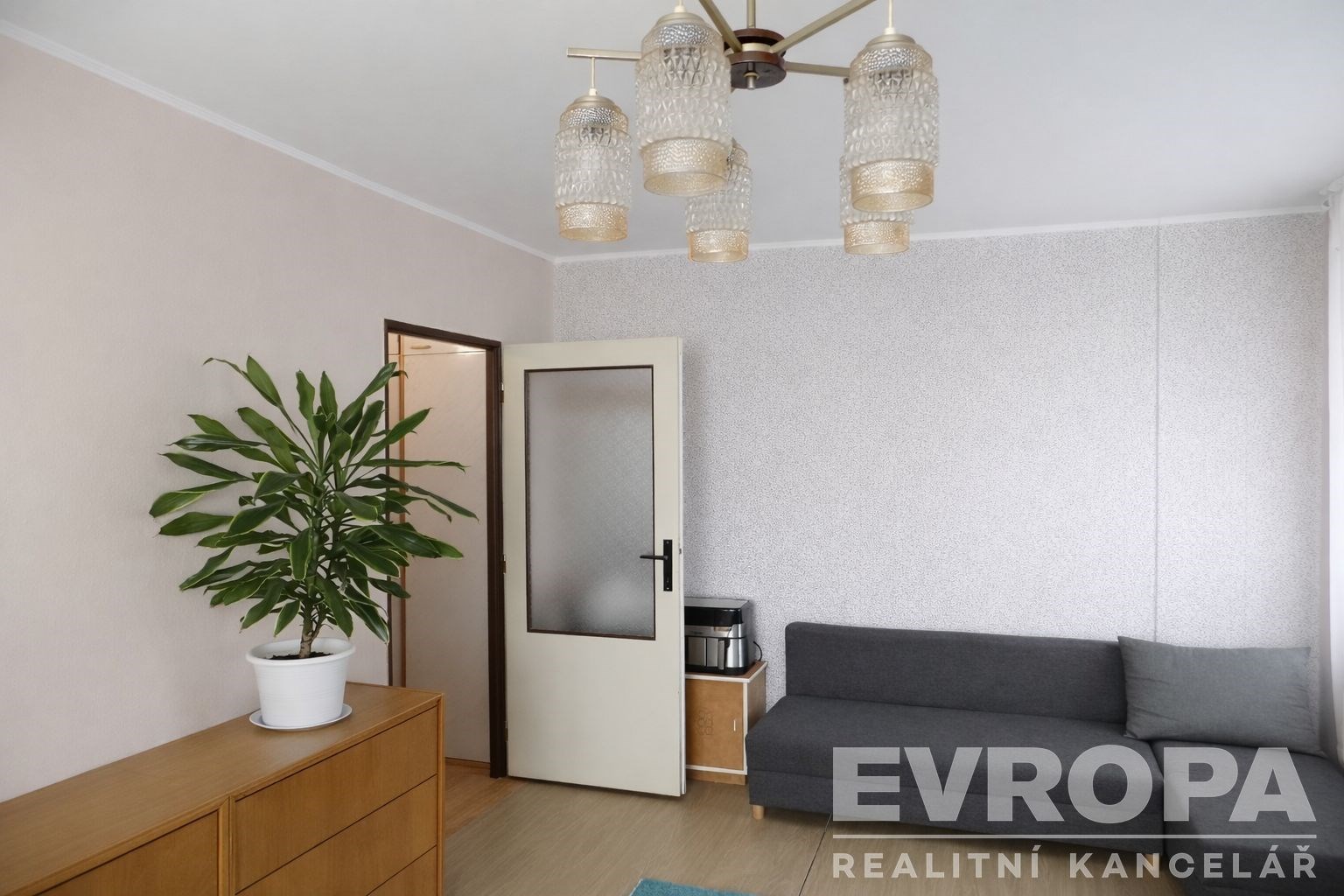 Světlý obývací pokoj s parketovou podlahou, moderní pohovka a designový lustr, interiér s neutrálními barvami a velkou rostlinou.