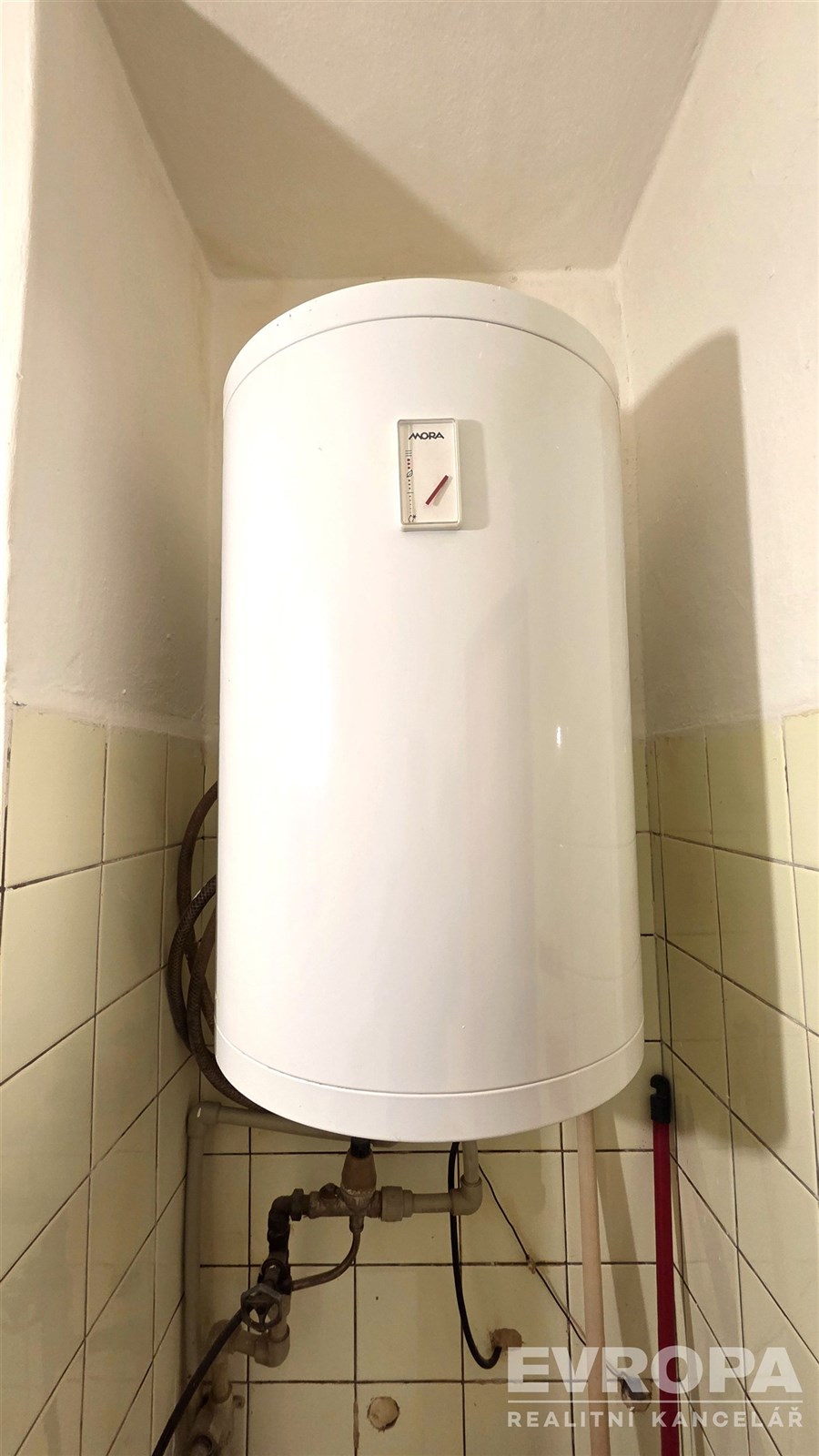 Elektrický bojler instalovaný v koupelně, obklopený bílými keramickými dlaždicemi, vhodný pro moderní interiéry a úsporný provoz.