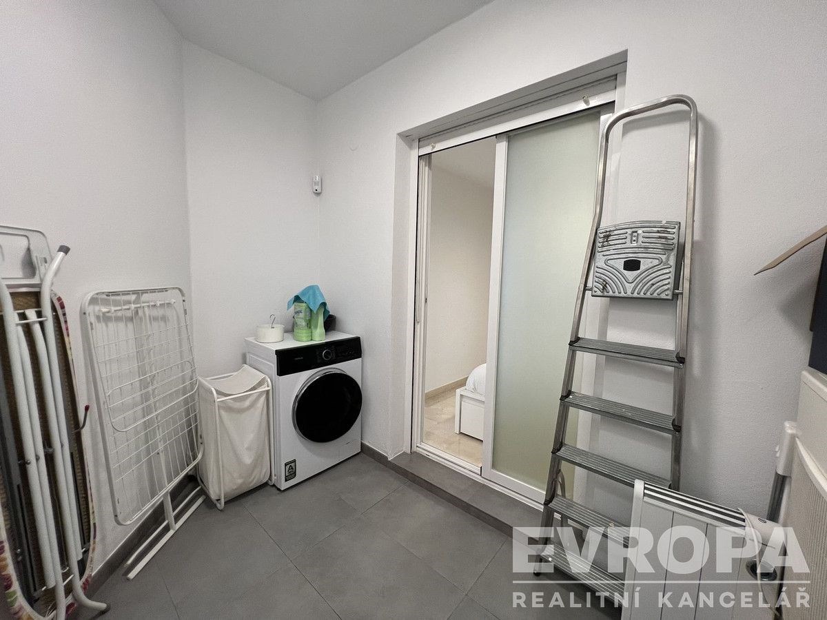 Prádelna s dlažbou, moderní pračka a sušák, světlé stěny, posuvné dveře oddělující prostor, praktické vybavení pro úklid.