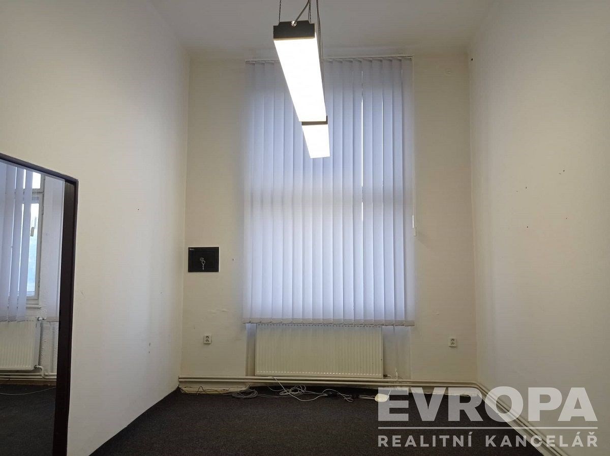 Prázdná místnost s vysokým stropem, kobercem a velkým oknem se žaluziemi, moderní závěsné osvětlení, bílé stěny.