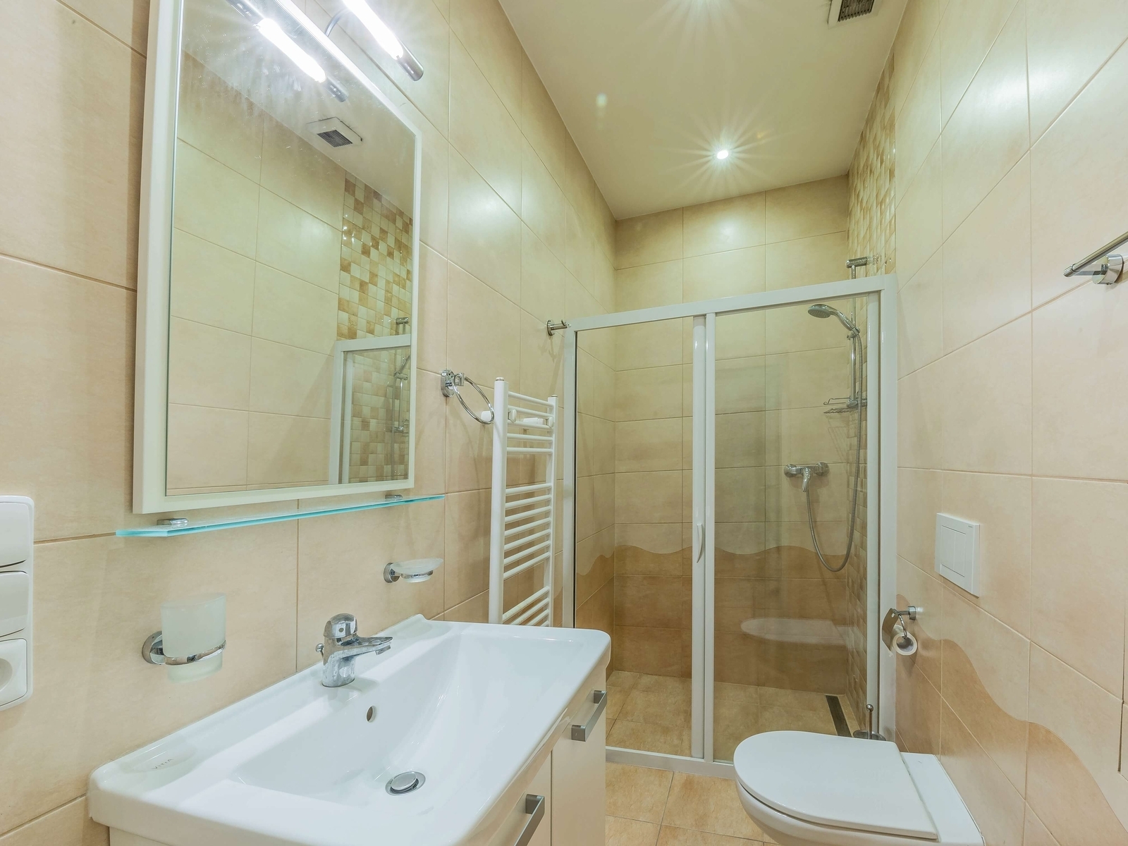 Druhá koupelna se sprchovým koutem, WC a topným žebříkem. Světlé obklady a bodová světla zajišťují čistý dojem.