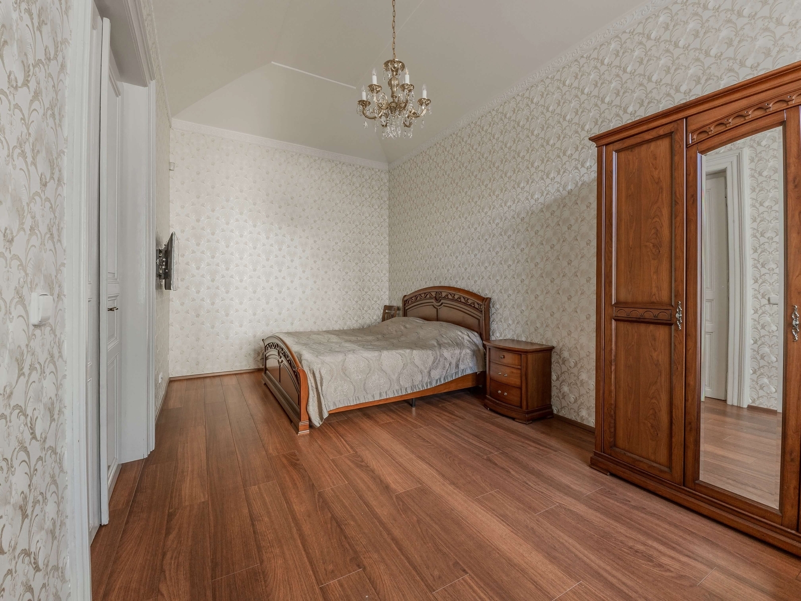 Prostorná ložnice s dřevěnou podlahou, šatní skříní a lustrem. Světlý interiér s vysokým stropem a klidnou atmosférou.
