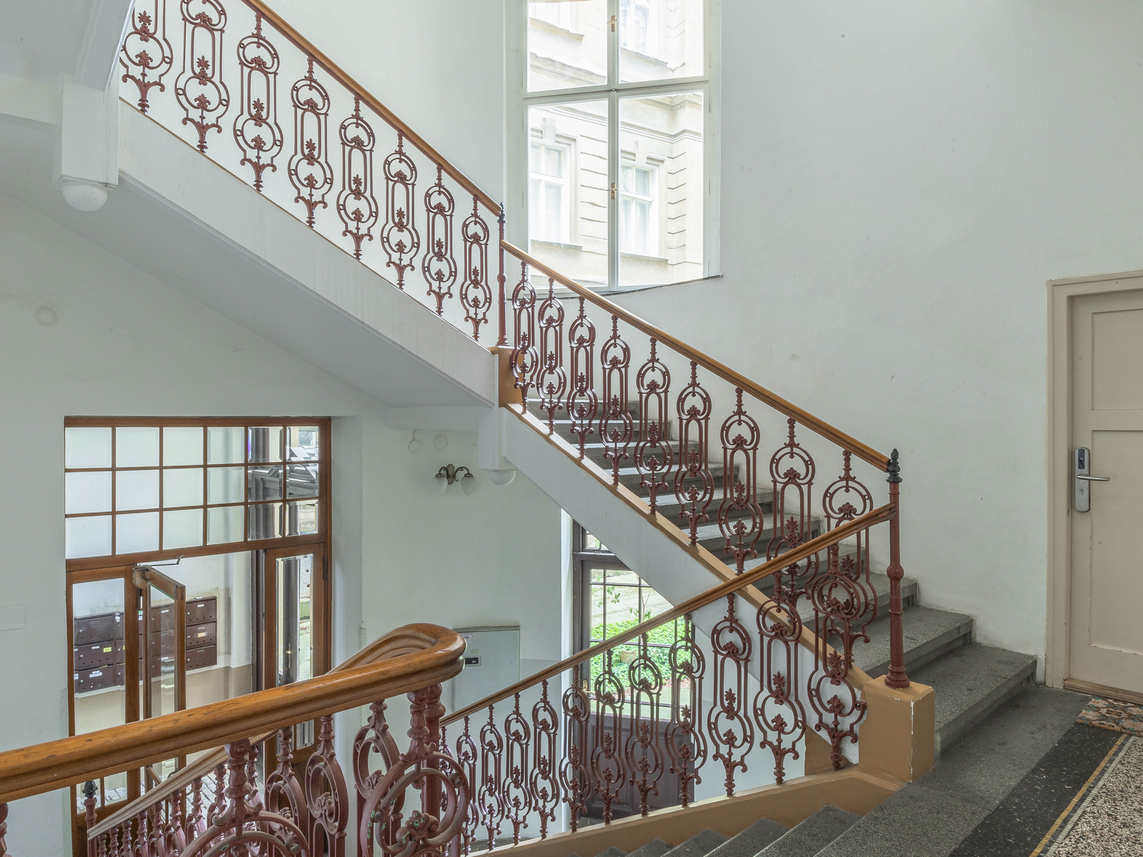 Historické schodiště domu s litinovým zábradlím a dřevěným madlem. Velká okna a vzdušné společné prostory.