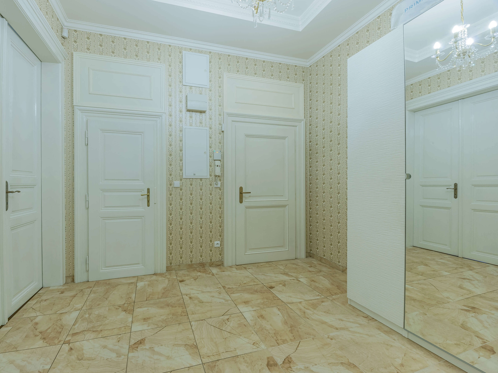 Široká vstupní hala s několika dveřmi a zrcadlovou skříní. Klasický vzhled, béžová dlažba a štukové lišty.