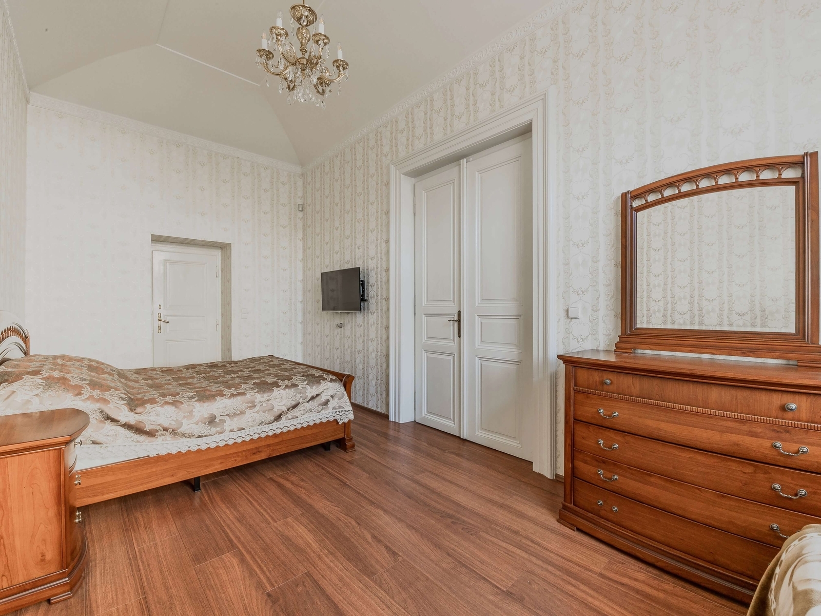 Ložnice s manželskou postelí, komodou a televizí u dveří. Dřevěná podlaha a vysoký strop podtrhují klid prostoru.