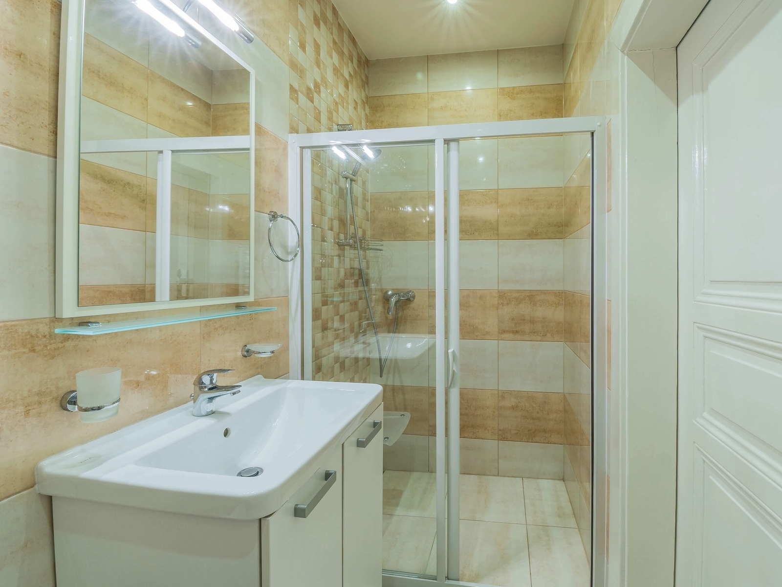 Moderní koupelna se sprchovým koutem a WC, velké zrcadlo nad umyvadlem. Světlé béžové obklady působí čistě.