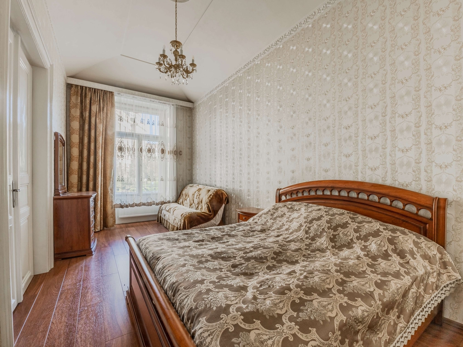 Světlá ložnice s manželskou postelí a toaletním stolkem. Velké okno do ulice, závěsy a lustr s křišťály.