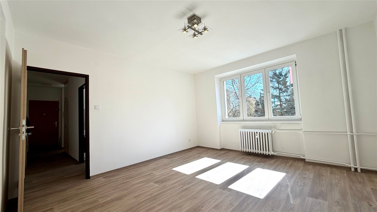 Světlý pokoj s parketovou podlahou, velkým oknem a moderním osvětlením, v rekonstruovaném bytě.