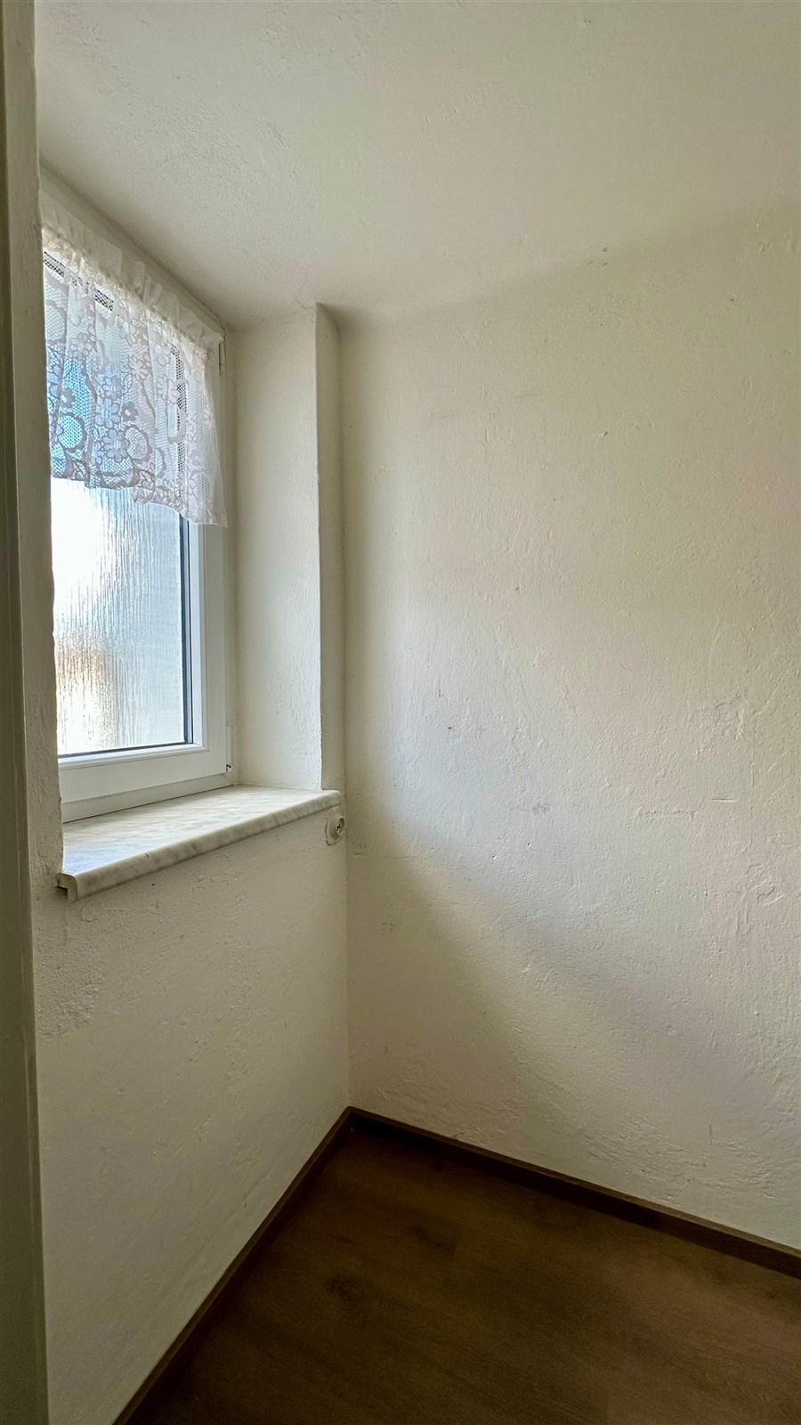 Malý pokoj s dřevěnou podlahou, omítnutými bílými zdmi a oknem s krajkovou záclonou, ideální pro skladování či kancelářský kout.