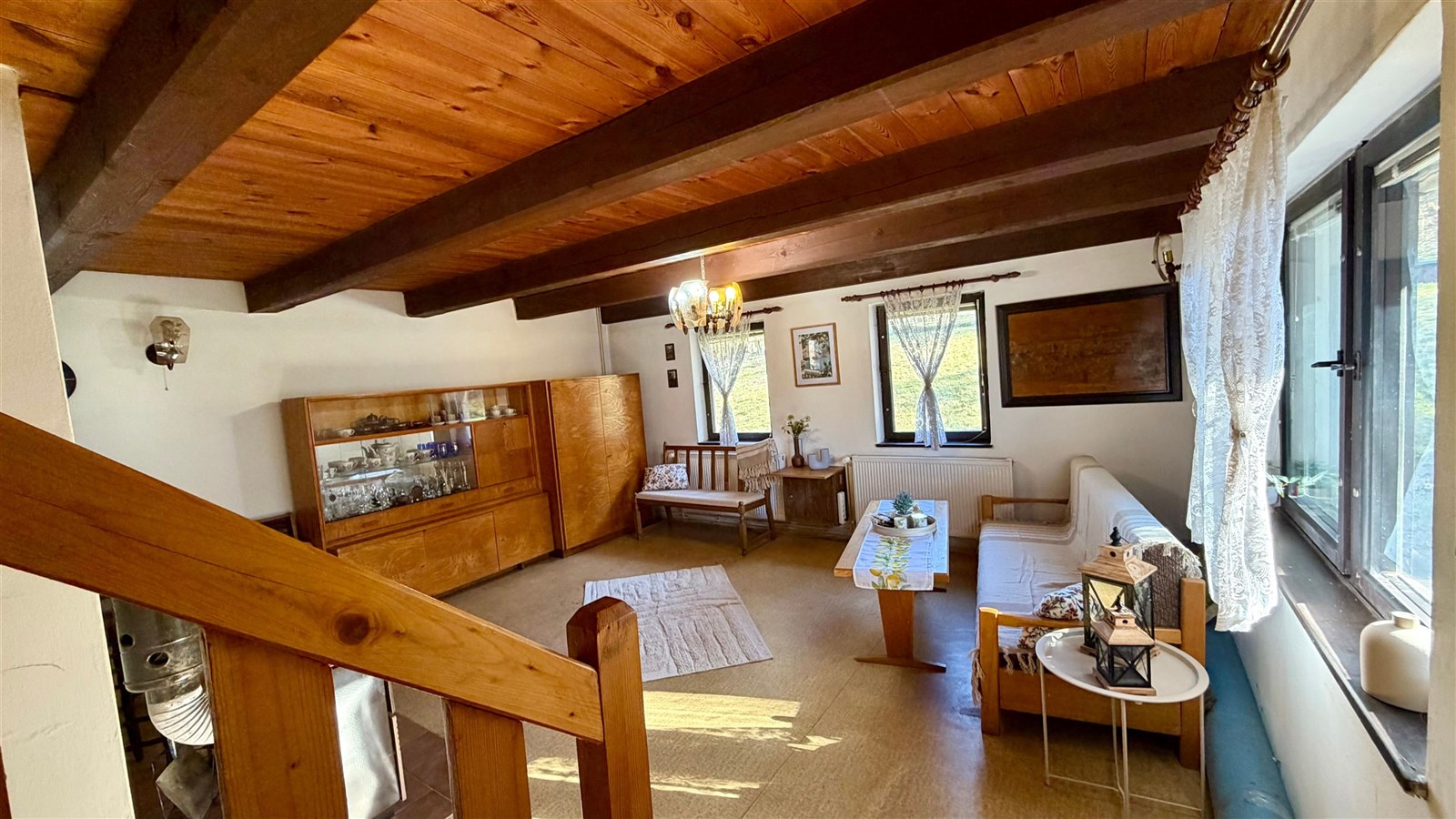 Obývací pokoj s dřevěným stropem, velkými okny a retro nábytkem. Světlá místnost, kde dřevo a světlo vytvářejí útulnou atmosféru.