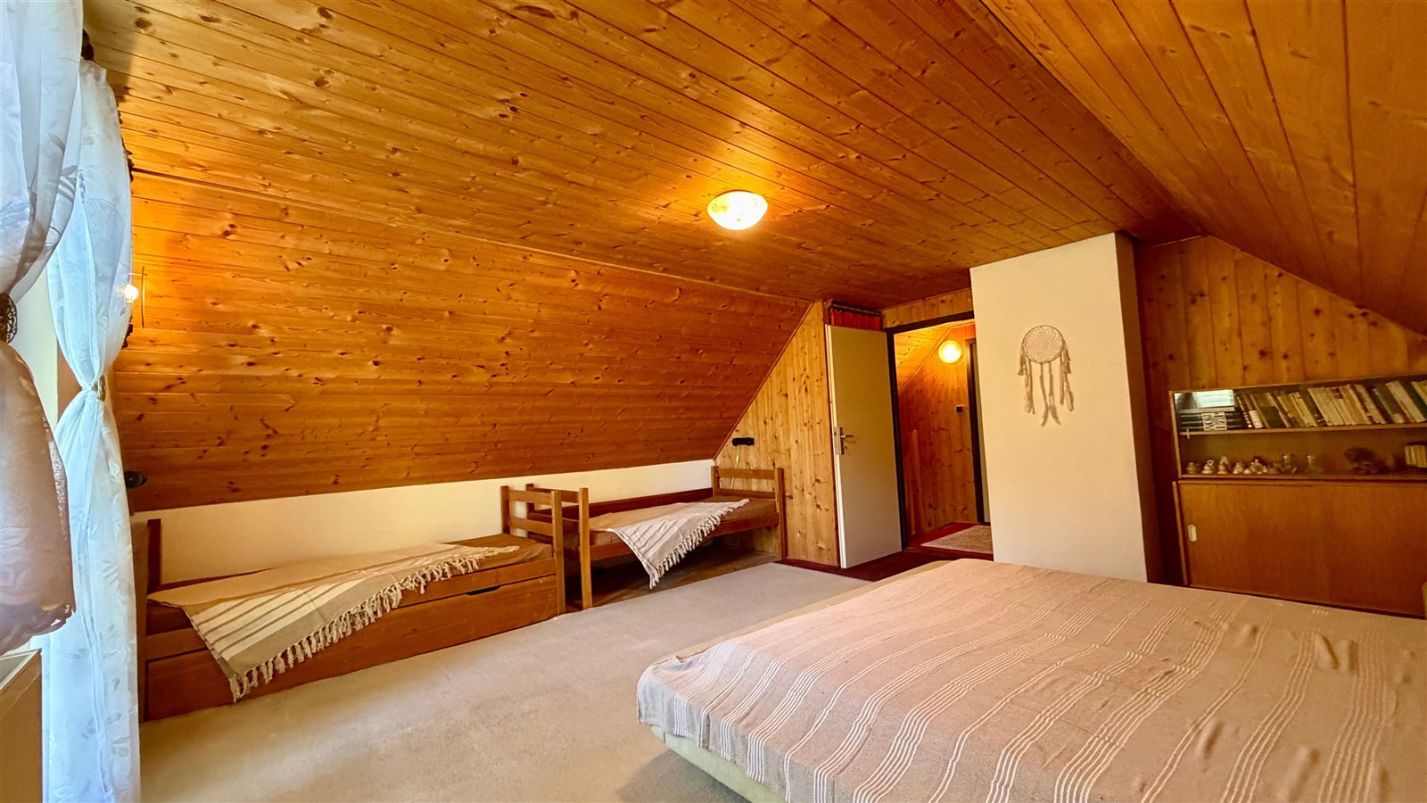 Podkrovní ložnice s dřevěným obložením, světlým kobercem a vestavěnými poličkami, nabízí útulný a rustikální styl bydlení.