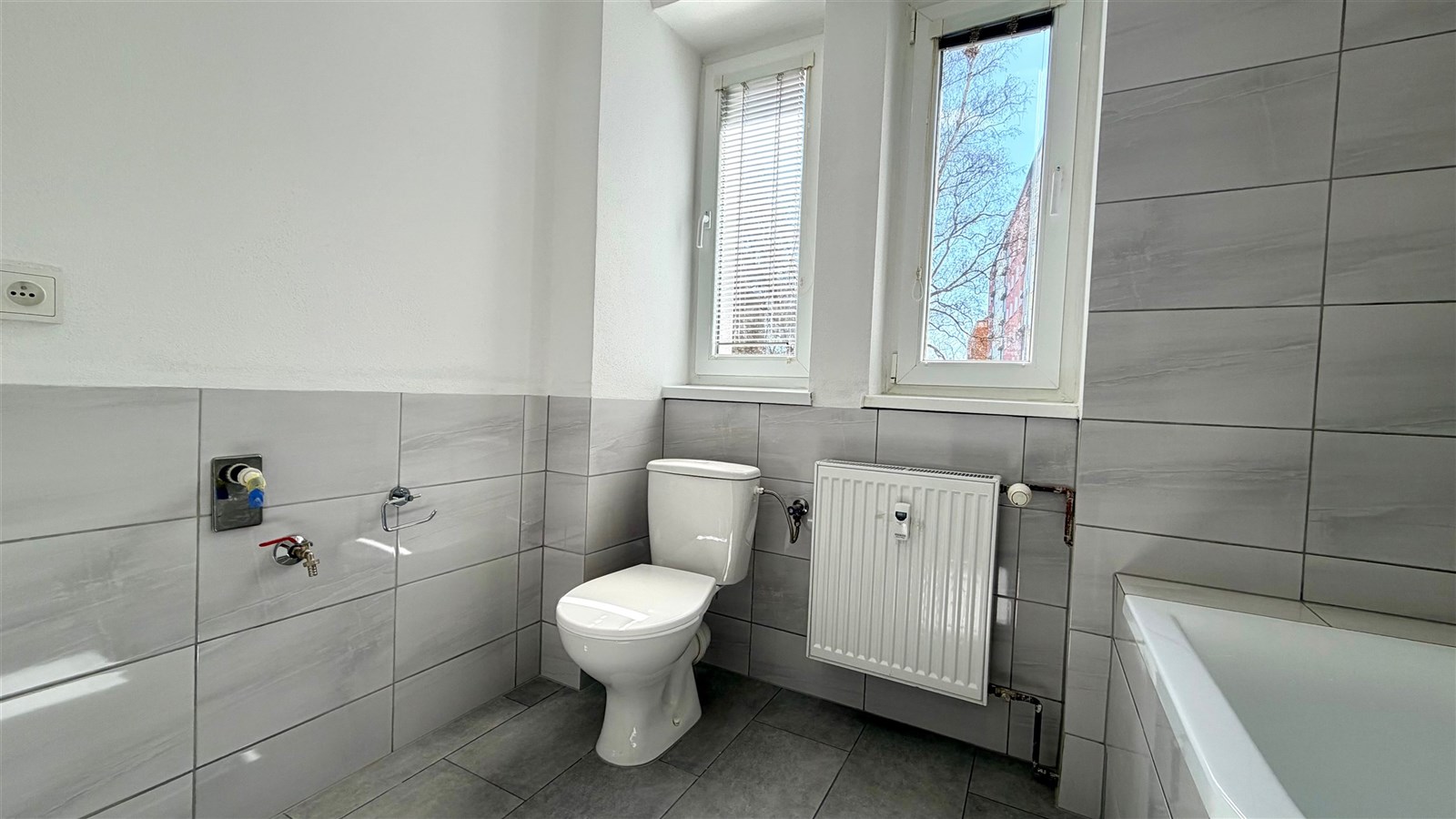 Moderní koupelna s šedou dlažbou, světlými obklady a velkým oknem, které zajišťuje dostatek přirozeného světla.