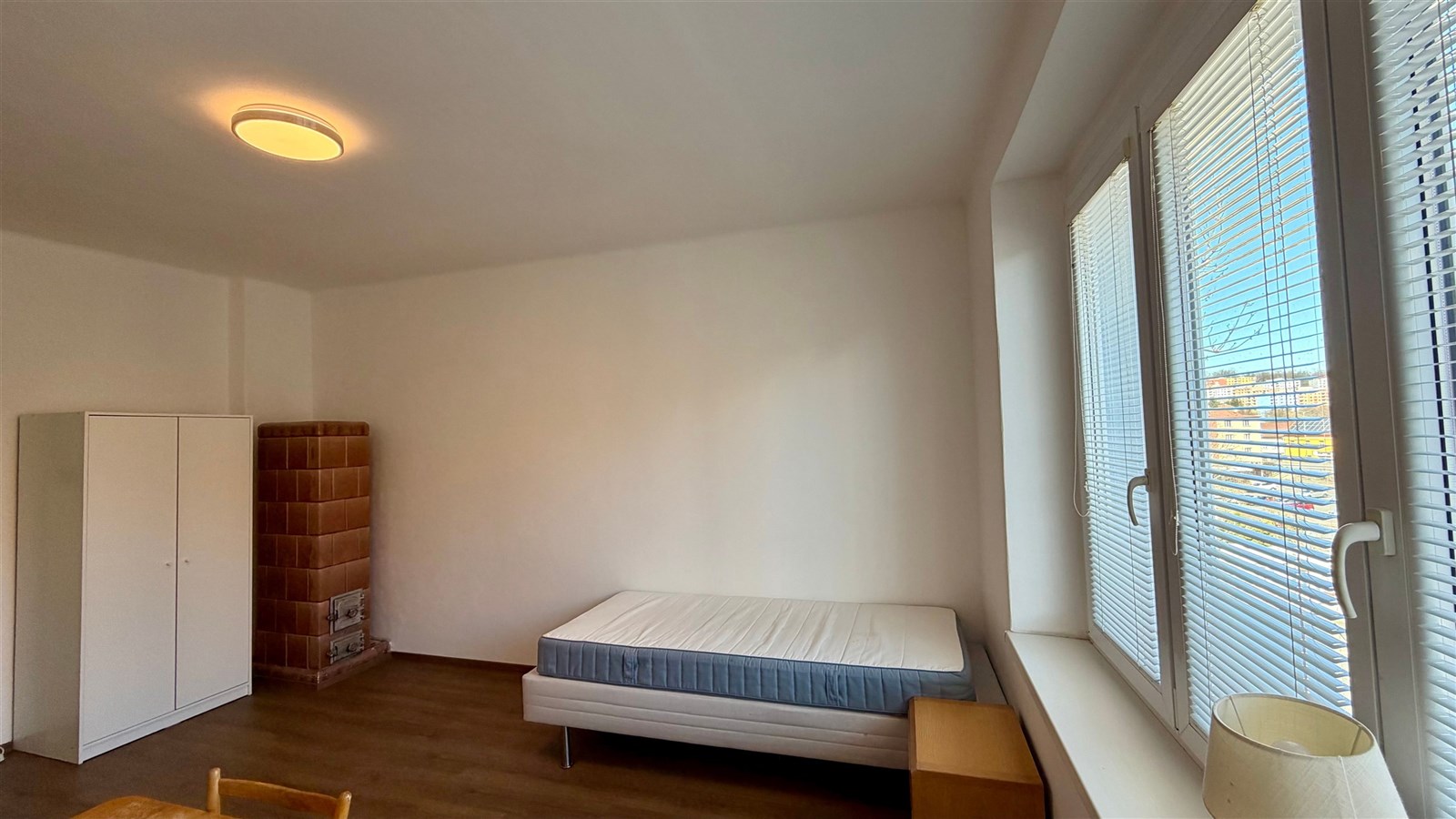 Světlý pokoj s parketovou podlahou, velká okna a vestavěné rolety, minimalistický styl, vhodný pro rekonstrukci či přizpůsobení dle potřeb.