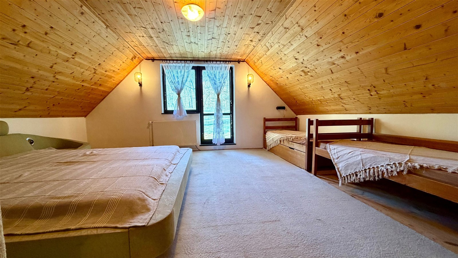 Podkrovní ložnice s dřevěným obložením, dvěma postelemi a kobercovou podlahou, světlá díky velkým oknům a modernímu osvětlení