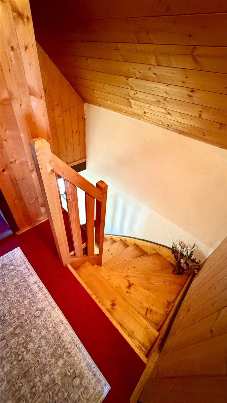 Dřevěné schodiště s červeným kobercem, obložené stěnami z přírodního dřeva, vede do podkroví, ideální pro rustikální interiér.