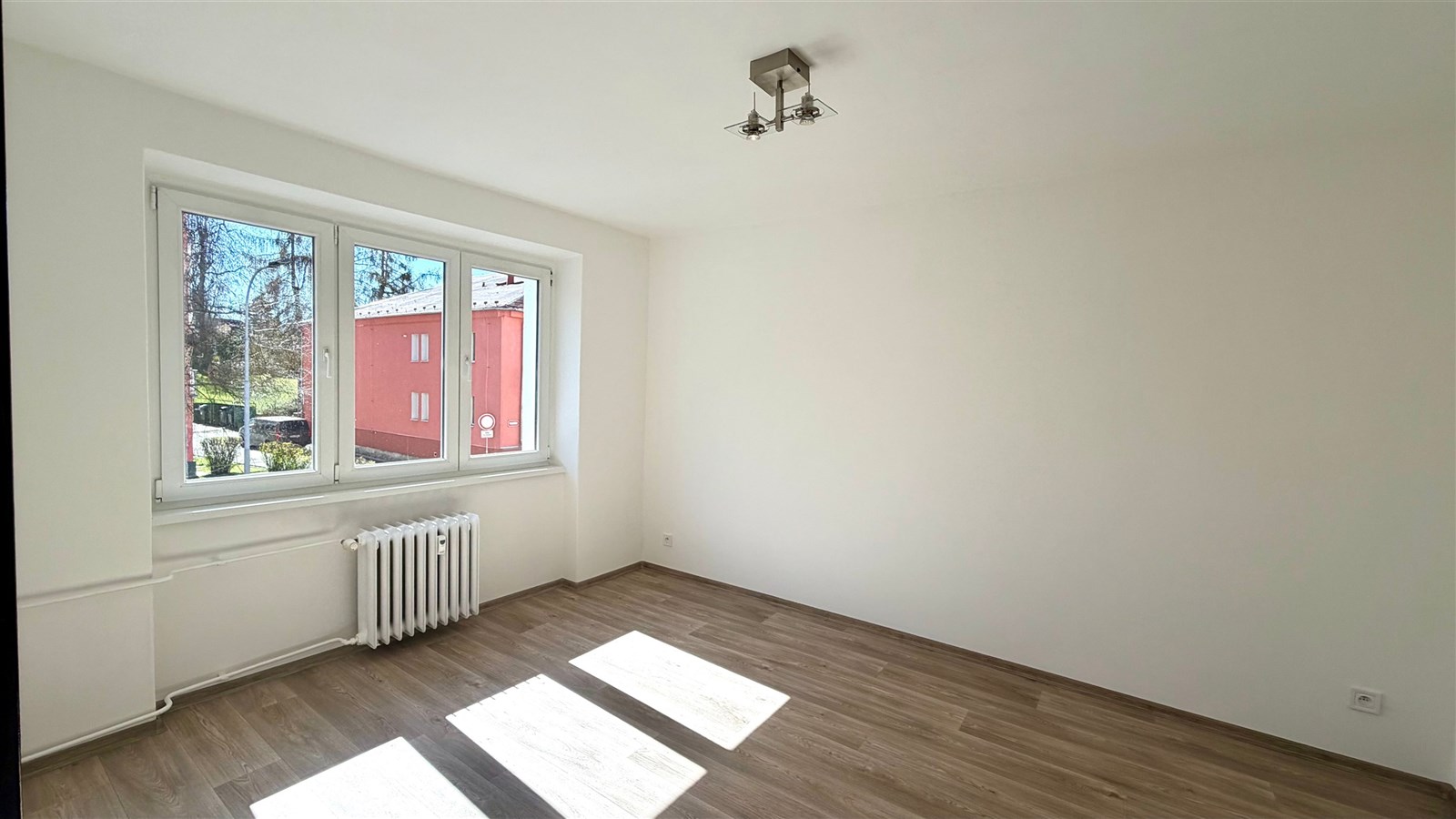 Světlý pokoj s moderními parketovými podlahami, velkými okny a rekonstruovanými omítkami, ideální pro útulné bydlení v městském prostředí.