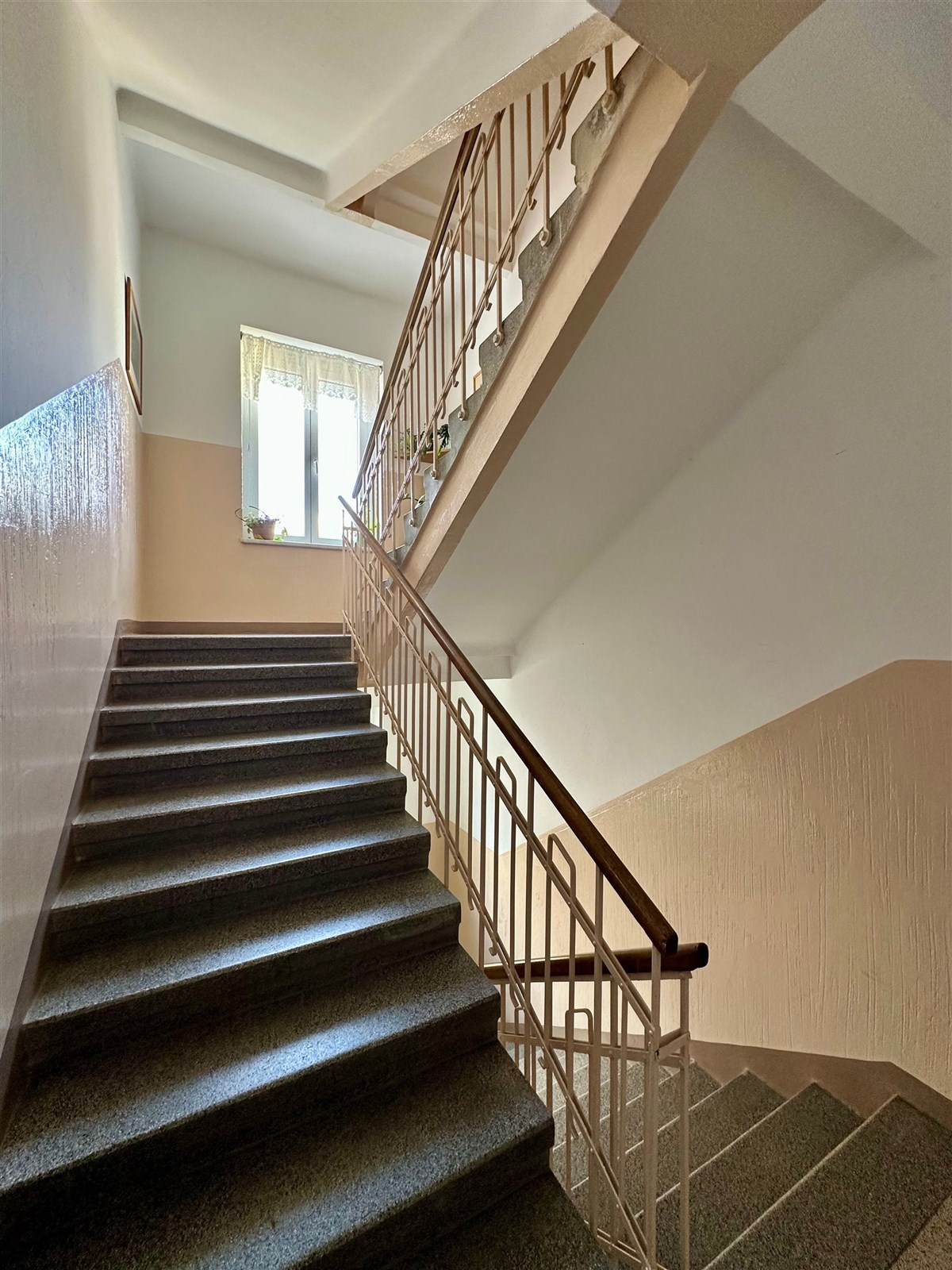 Schodiště s kamennými schody, velkým oknem a zábradlím v retro stylu, nabízí prostorný a světlý průchod v dobře udržovaném bytovém domě.