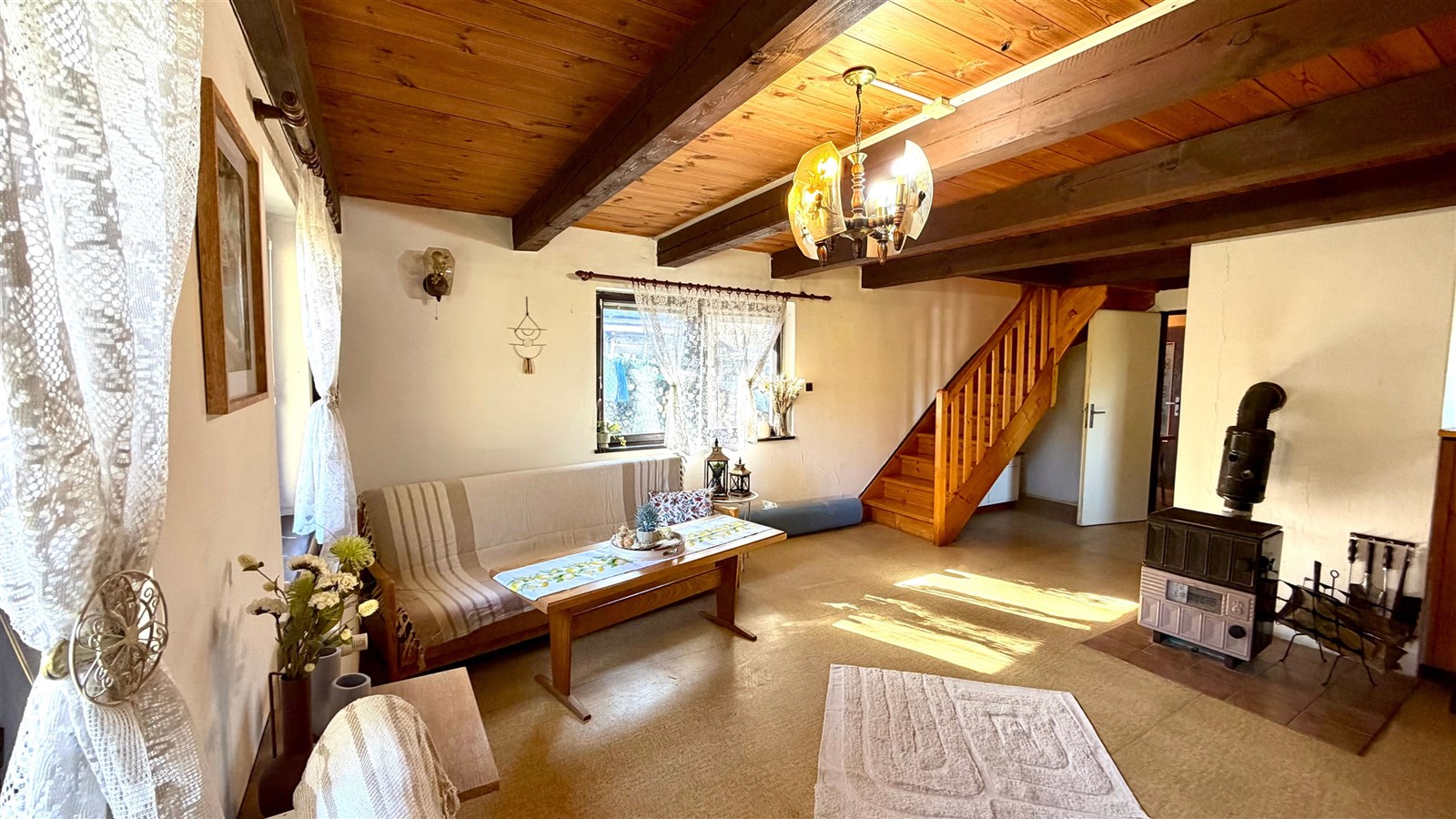 Světlý obývací pokoj s dřevěným stropem, krbovými kamny a velkými okny. Interiér v retro stylu nabízí útulnou atmosféru a praktické uspořádání.