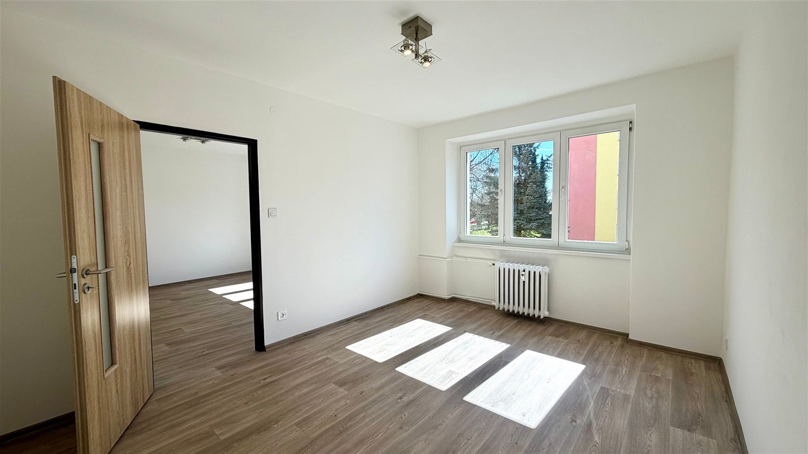 Světlý pokoj s moderní parketovou podlahou, velkým oknem a novým osvětlením.