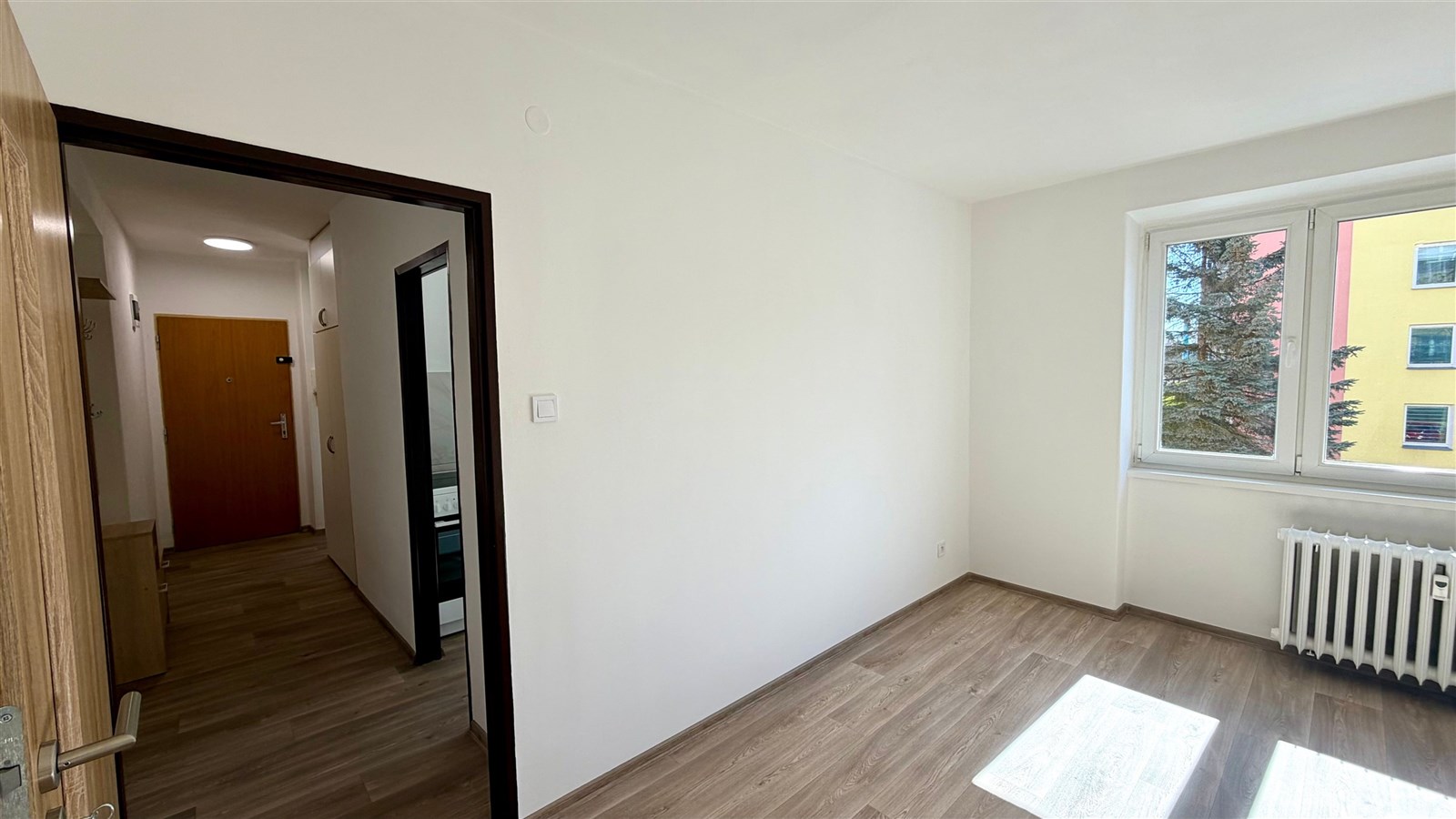 Světlý pokoj s parketovou podlahou, velkým oknem a moderním bílým radiátorem, sousedí s chodbou s dřevěnými dveřmi.