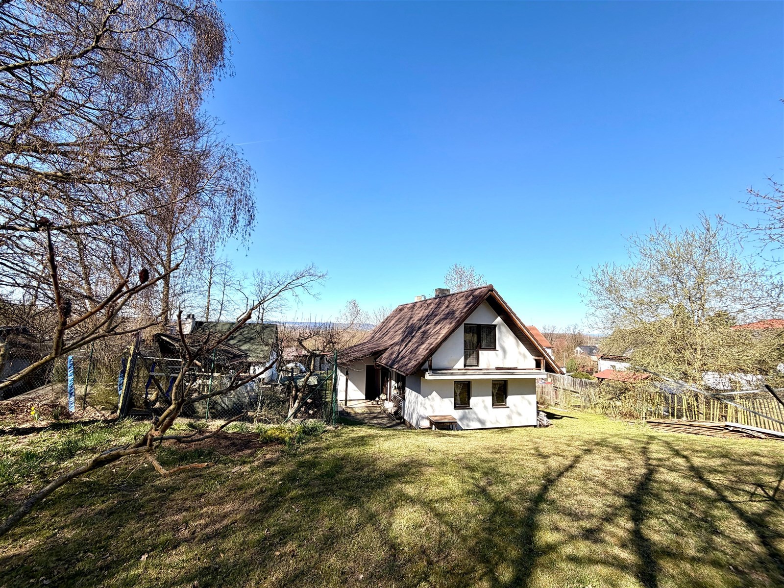 Dům s šikmou střechou a bílou fasádou na prostorném pozemku, obklopen stromy a trávníkem, v klidné venkovské lokalitě vhodný k rekreaci