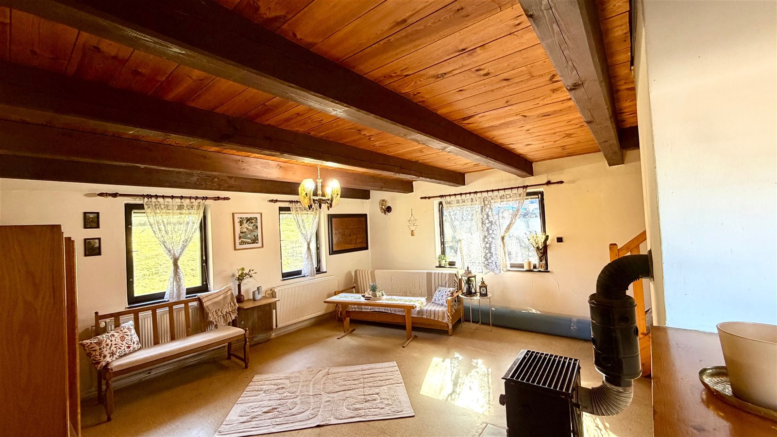 Prostorný obývací pokoj s dřevěným stropem, velká okna, krbová kamna, rustikální styl, světlý a útulný prostor pro relaxaci.