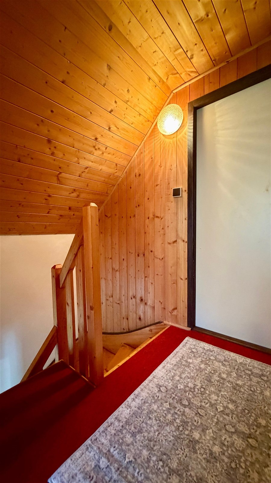 Chodba s dřevěným obkladem, červeným kobercem a moderním osvětlením nabízí útulný prostor ve stylu chalupy.