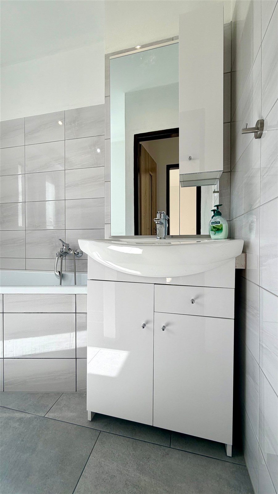 Moderní koupelna s dlažbou, bílým umyvadlem a zabudovanou skříňkou, světlé dlaždice, elegantní design, prostorná a světlá.