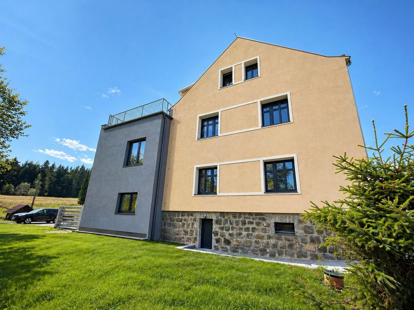 Třípatrový rodinný dům s moderními fasádními prvky a velkými okny, situovaný v klidné lokalitě obklopené zelení, s dobře udržovaným trávníkem.