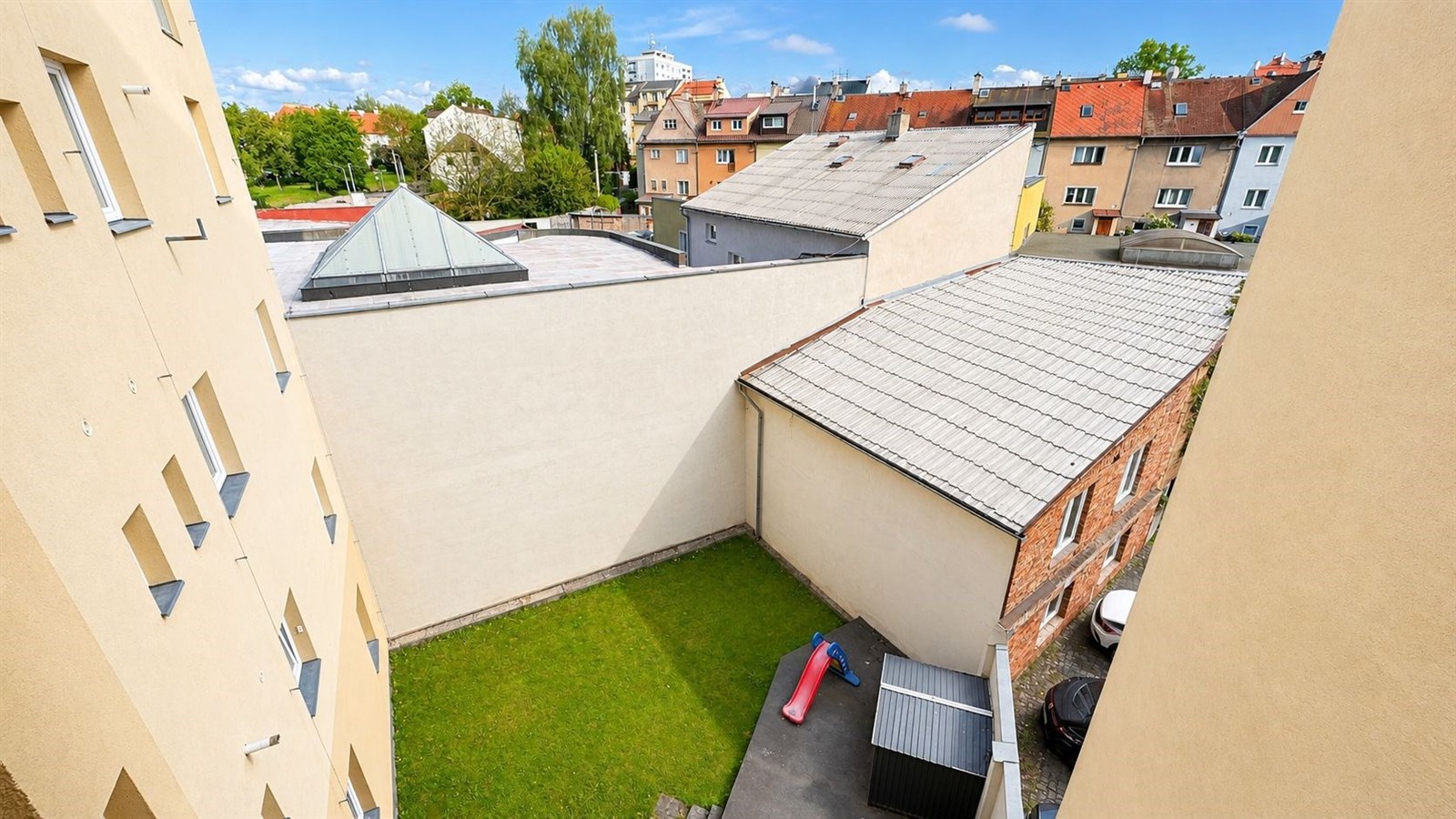 Pohled z okna na dvůr s trávníkem a okolními budovami, střešní krytina a cihlové zdi, klidná lokalita.